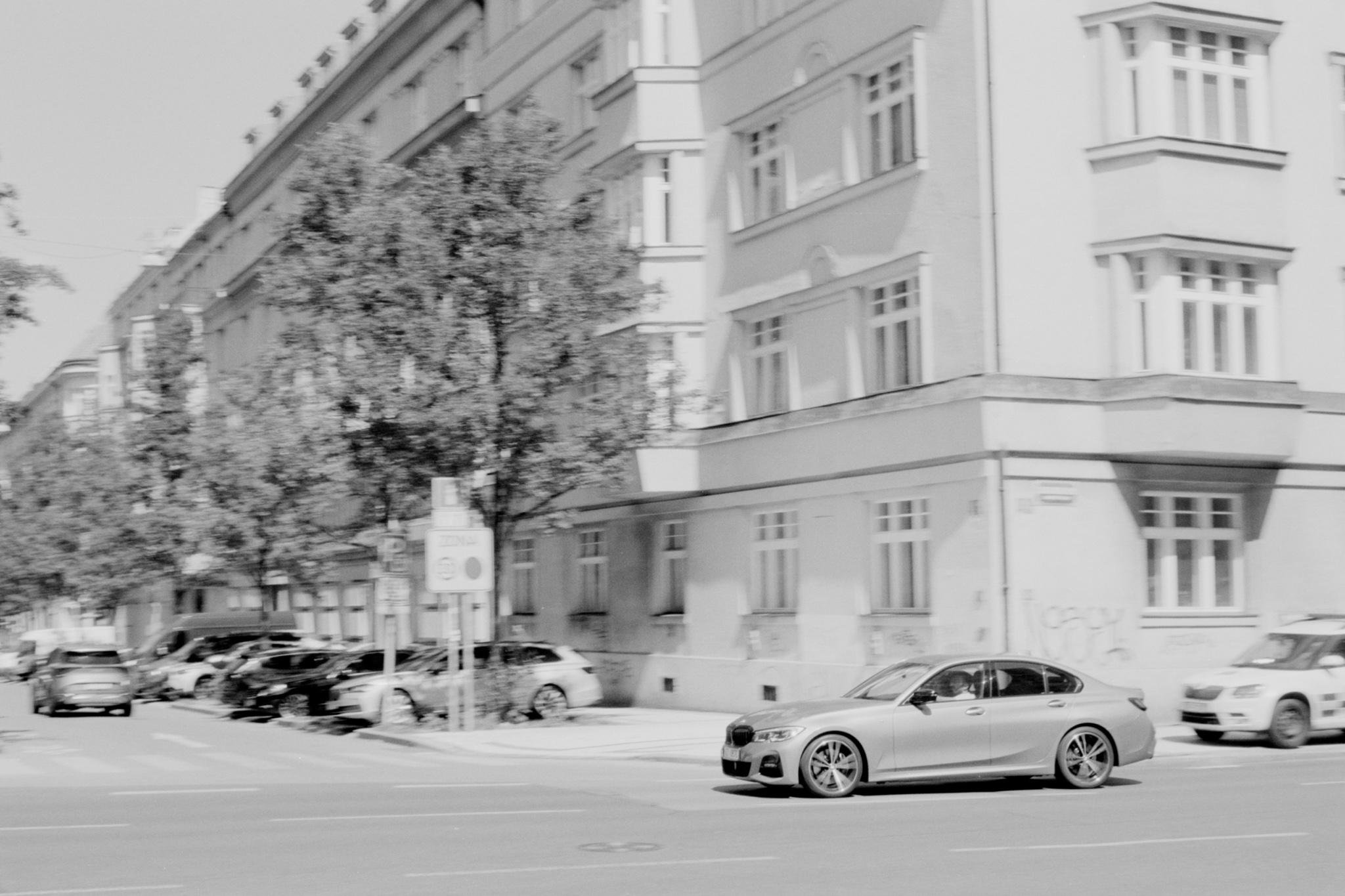Black-and-white street scene with a silver sedan driving past a corner apartment building with large windows; parked cars line the curb beneath leafy trees, viewed from across a wide intersection.