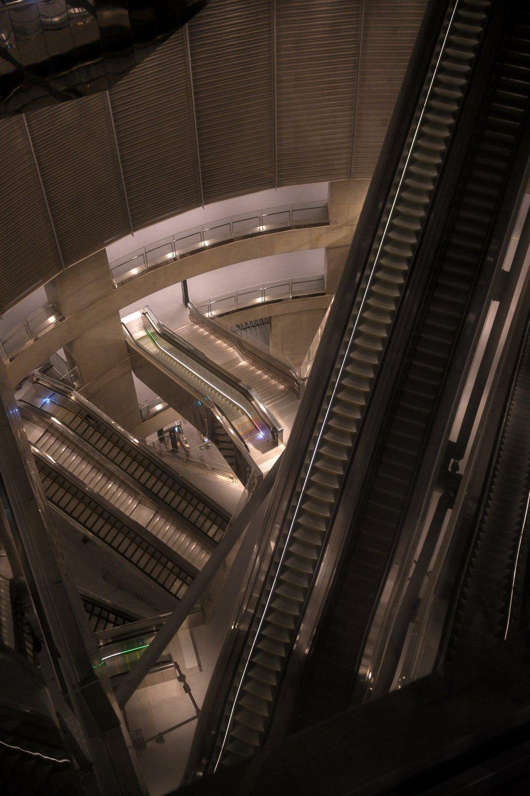 Photo verticale. Plongée sur un lieu très profond, des escalators barrent la vue dans de multiples directions