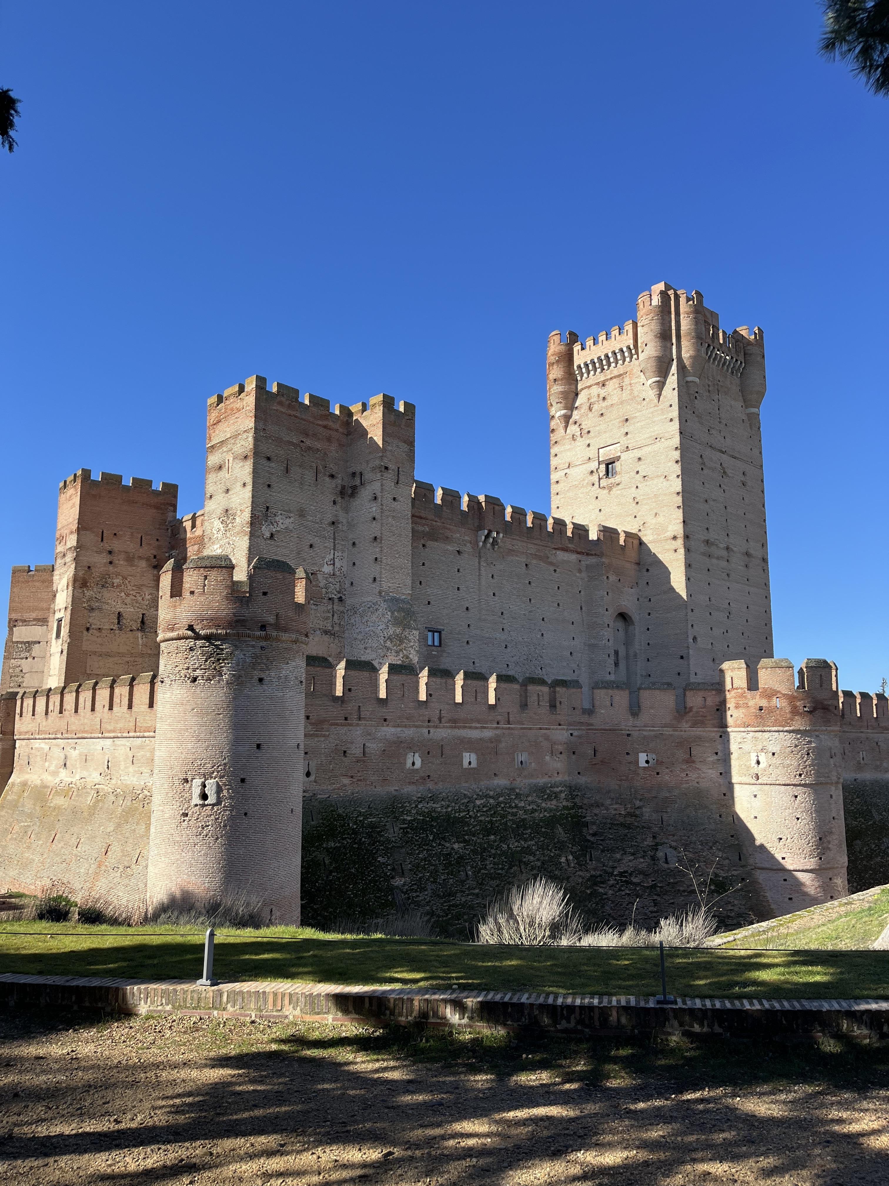 This image features a large, well-preserved medieval castle. It has fortified walls, battlements, and a tall central tower. The structure appears to be made of stone and brick, with architectural features typical of medieval fortifications, such as crenellated parapets and circular towers. The sky is clear and blue, suggesting it was taken on a sunny day.