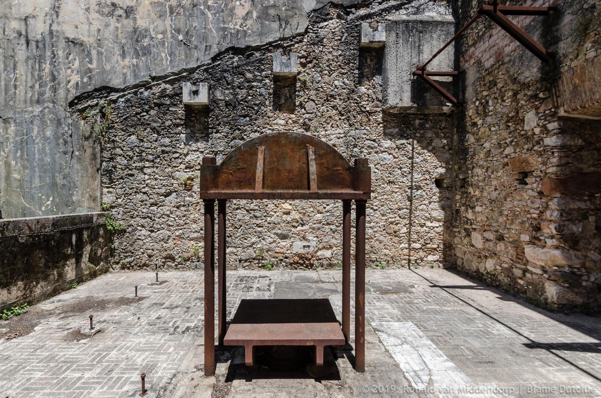 Rusty metal frame with a rounded top standing on a concrete floor. Set inside a walled stone-and-concrete courtyard with exposed beams and scattered floor bolts.