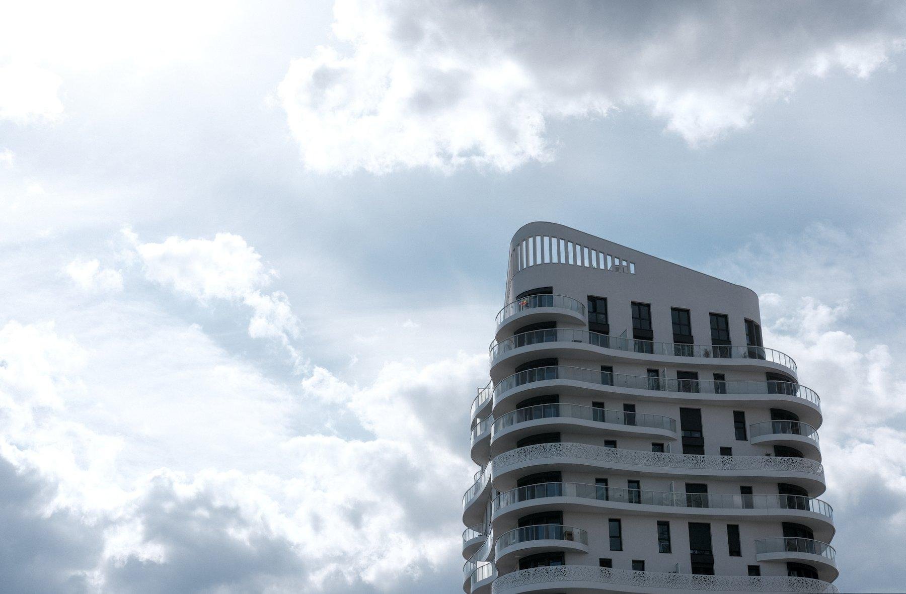 Photo horizontale. Sur la droite, le haut d’un immeuble aux formes arrondies très moderne, sous un ciel bleu mais très chargé en nuage. Un halo de lumière arrive du coin haut gauche