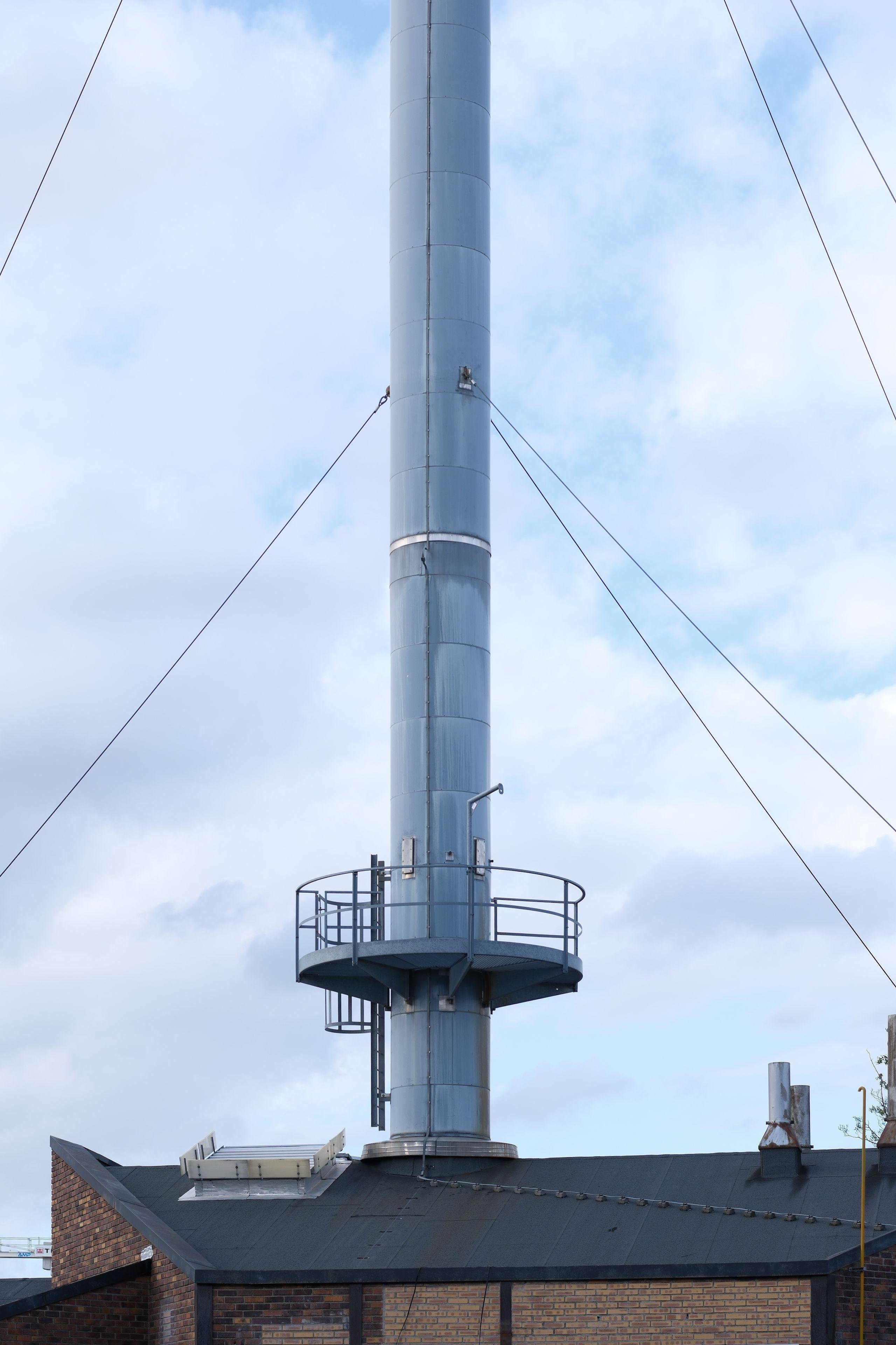 Photo verticale. Une partie de cheminée en métal (je crois) avec une plateforme de maintenance circulaire quelques mètres au dessus du toit d’un bâtiment. Des câbles tendus stabilisent la cheminée