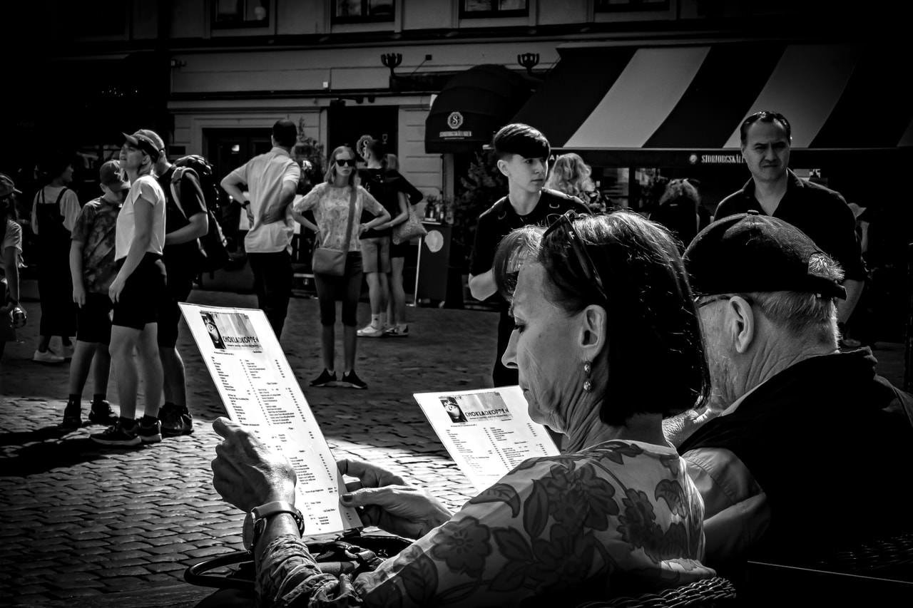 Two people sitting outdoors, holding and reading menus in a cobblestone street setting. In the background, several pedestrians, including a group standing and a couple of individuals walking, appear in front of a building with a striped awning. The image is in black and white.