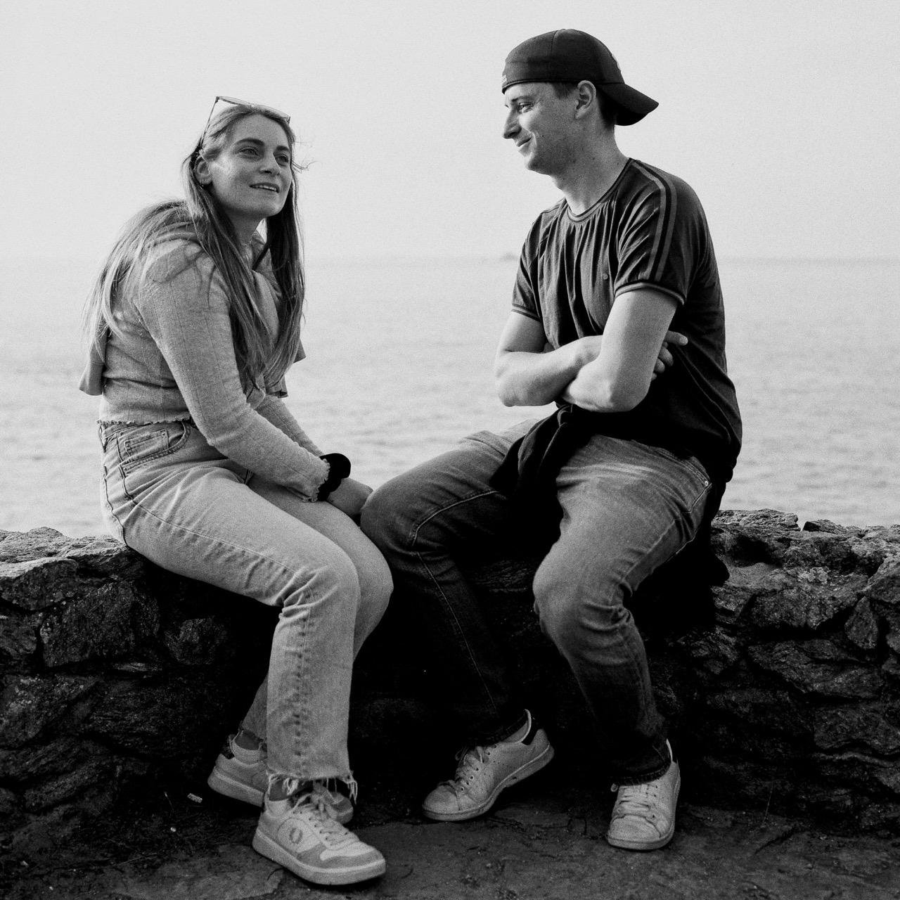 The image shows a black and white photograph of two people sitting on a stone wall. They are by the water, possibly on a sea or lakeside. The person on the left is wearing jeans and sneakers, while the person on the right is wearing a t-shirt, jeans, and a backwards cap. Both appear to be engaged in a conversation or interaction.