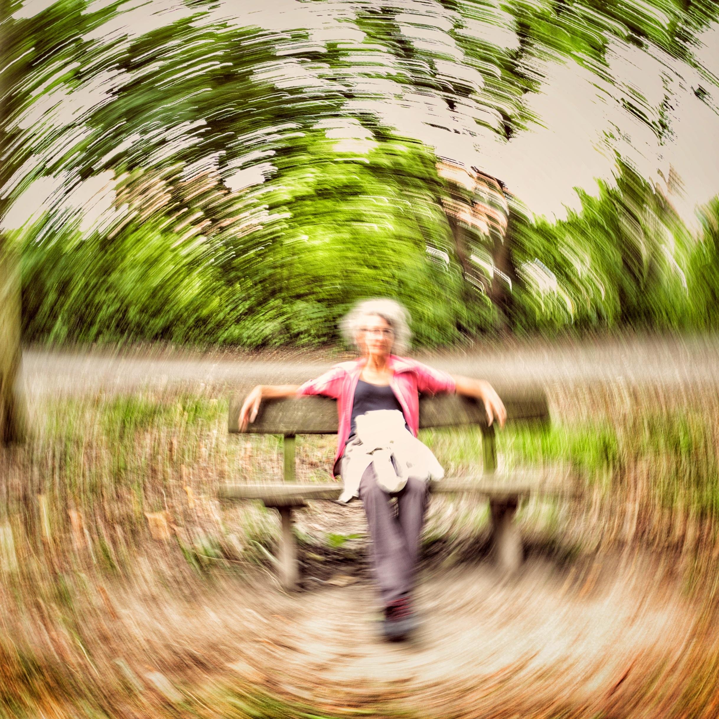 Person sitting on a wooden bench in a park, arms stretched along the backrest, with green trees and a path behind; strong circular motion blur radiates around the subject. ### 
Eine Person sitzt auf einer Holzbank in einem Park, die Arme entlang der Rückenlehne ausgestreckt, im Hintergrund grüne Bäume und ein Weg; eine starke kreisförmige Bewegungsunschärfe strahlt um das Motiv herum aus.