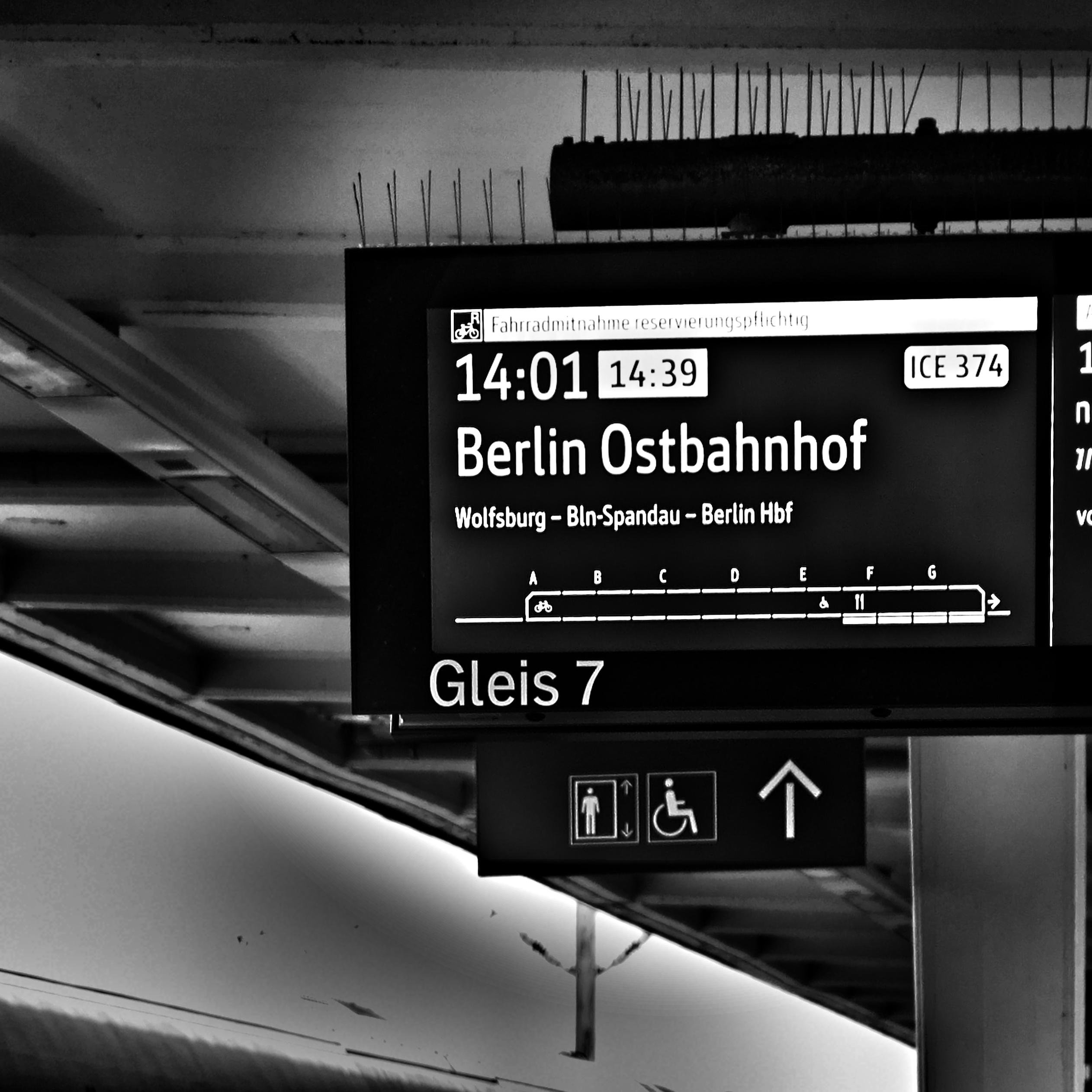 Electronic train platform sign under a station roof reading “Berlin Ostbahnhof,” showing times 14:01 and 14:39, train ICE 374, route “Wolfsburg – Bln-Spandau – Berlin Hbf,” and “Gleis 7,” with icons for restrooms, wheelchair access, and an upward arrow. 
### 
Elektronische Bahnsteiganzeige unter einem Bahnhofsdach mit der Aufschrift „berlin ostbahnhof“, den Uhrzeiten 14:01 und 14:39, dem Zug ICE 374, der Route „Wolfsburg – Bln-Spandau – Berlin Hbf“ und „Gleis 7“, sowie Symbolen für Toiletten, Rollstuhlzugang und einem Pfeil nach oben.
