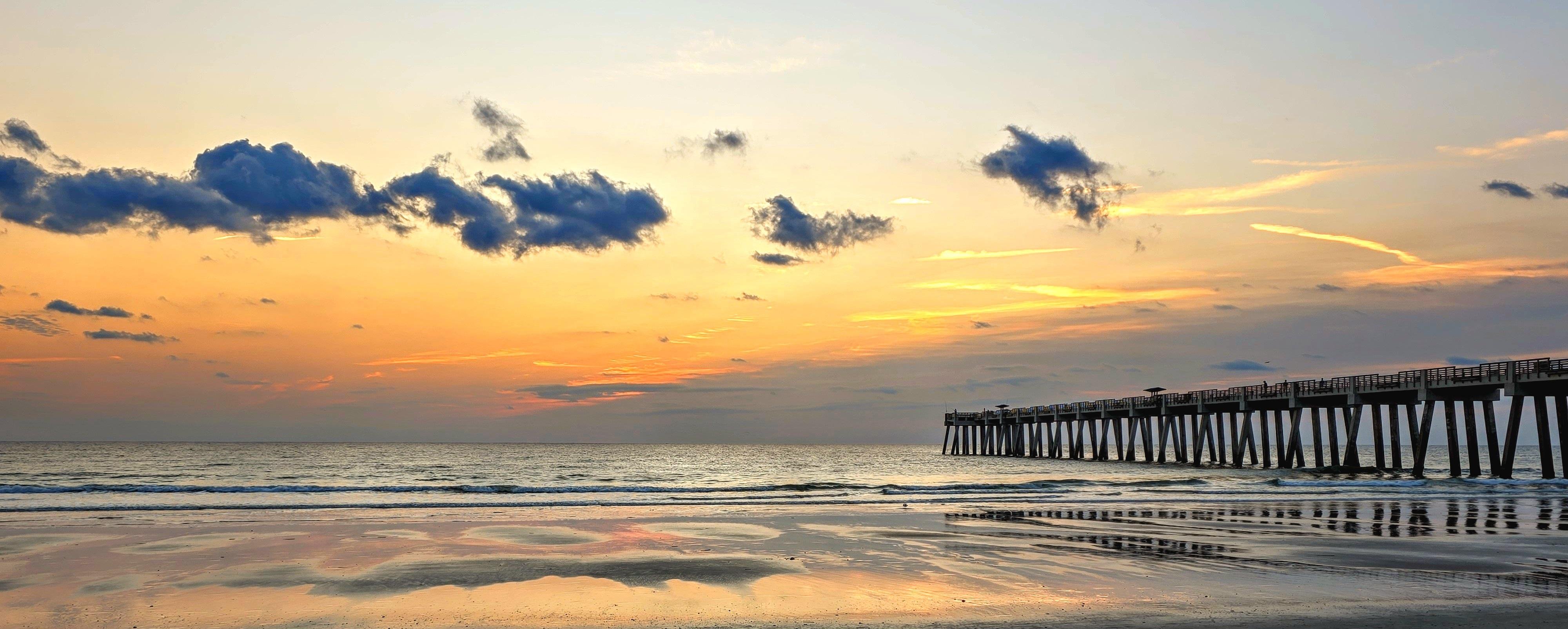 Sunrise over a beach with a pier extending into the ocean on the right. The sky displays shades of orange, yellow, and blue with scattered clouds. The wet sand reflects the colors of the sky, adding to the serene atmosphere.
