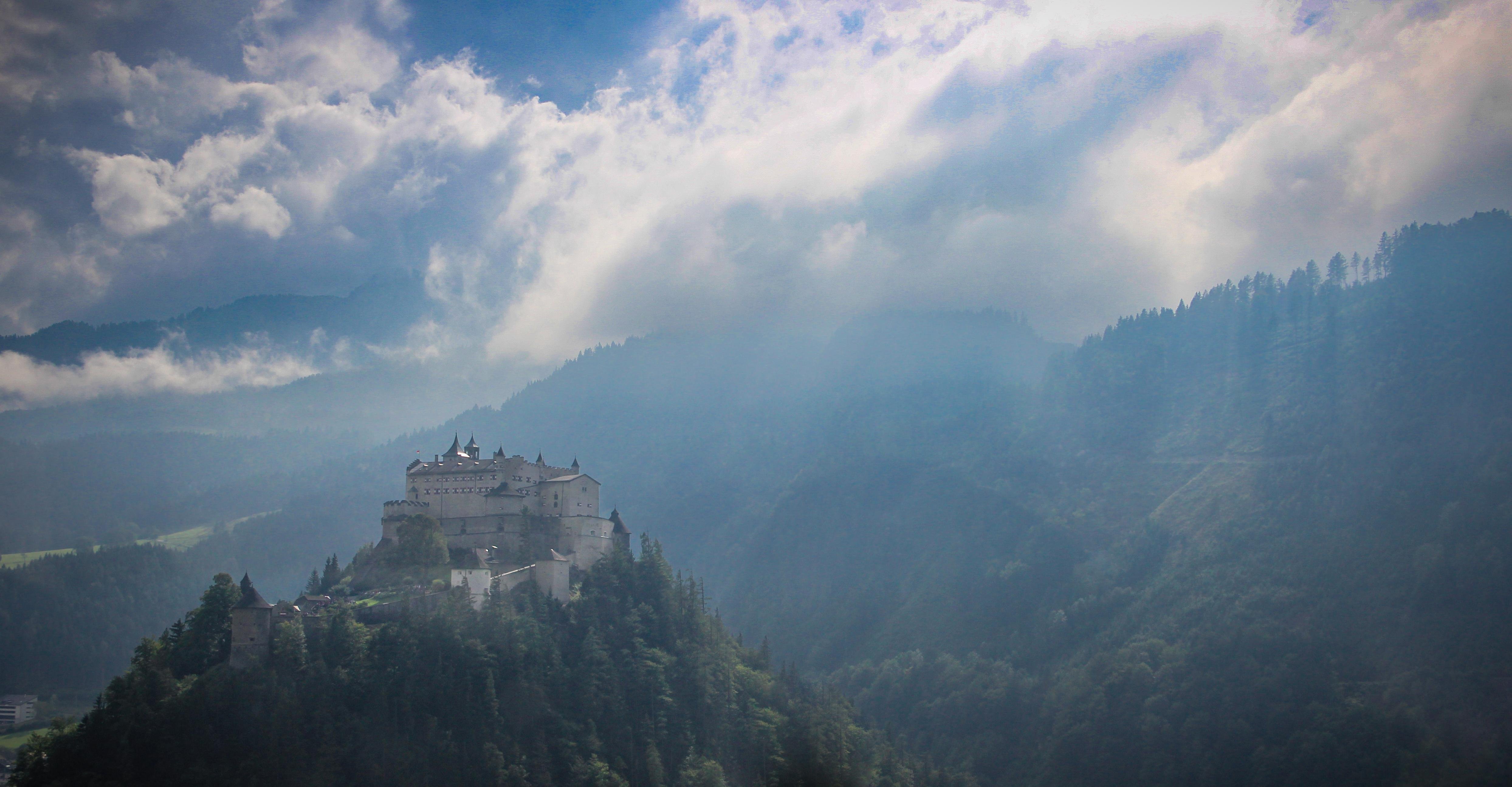 The image shows a majestic castle set atop a hill surrounded by lush green forests. In the background, there are mountains partially obscured by clouds and mist, creating a dramatic and ethereal atmosphere. The sunlight piercing through the clouds adds a mystical quality to the scene.
