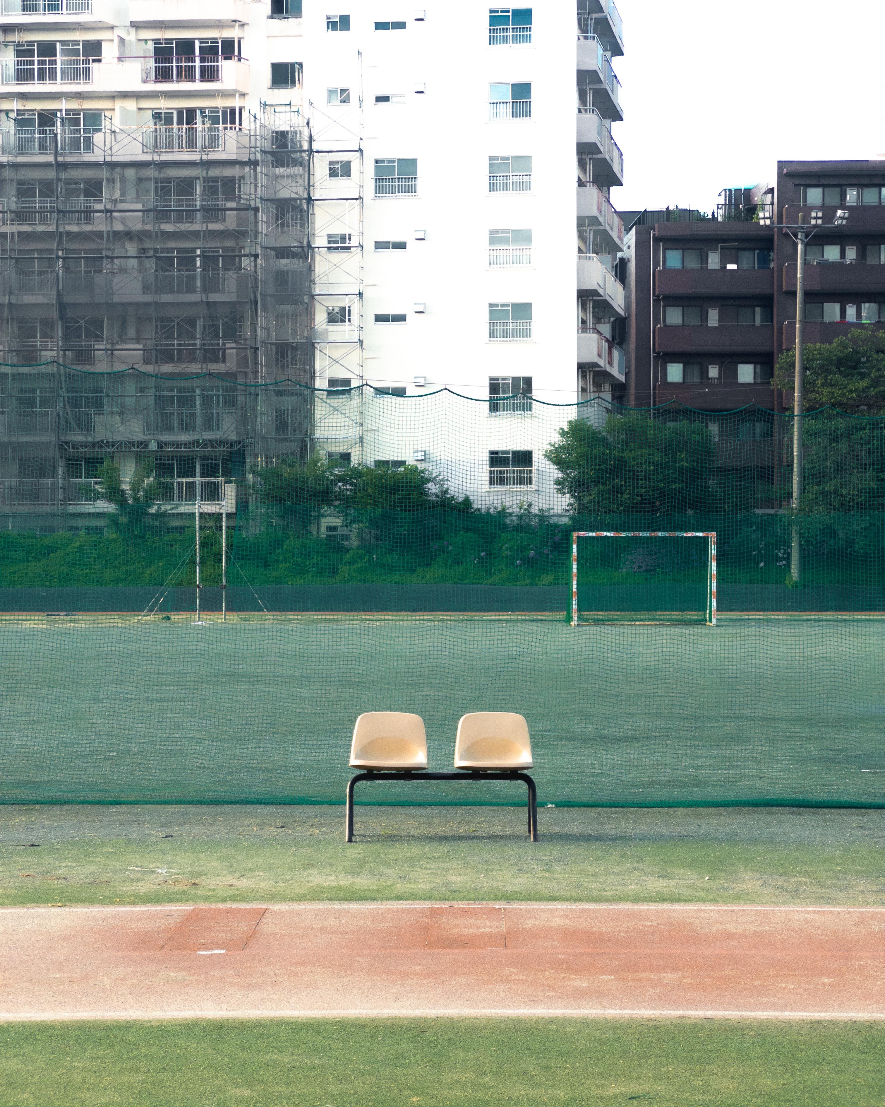 Two cream-colored chairs on a metal frame positioned on a track, facing a fenced soccer field. The field has a goalpost and is partially enclosed with netting. In the background, there are high-rise buildings, some with scaffolding, surrounded by greenery.