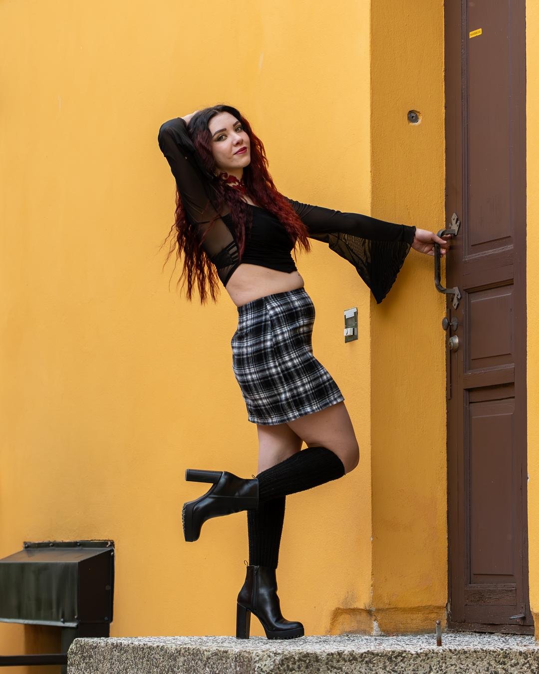 The image shows a person posing in front of a yellow wall. They are wearing a black crop top with flared sleeves, a plaid skirt, and black knee-high boots. They are standing near a brown door, leaning with one foot raised.