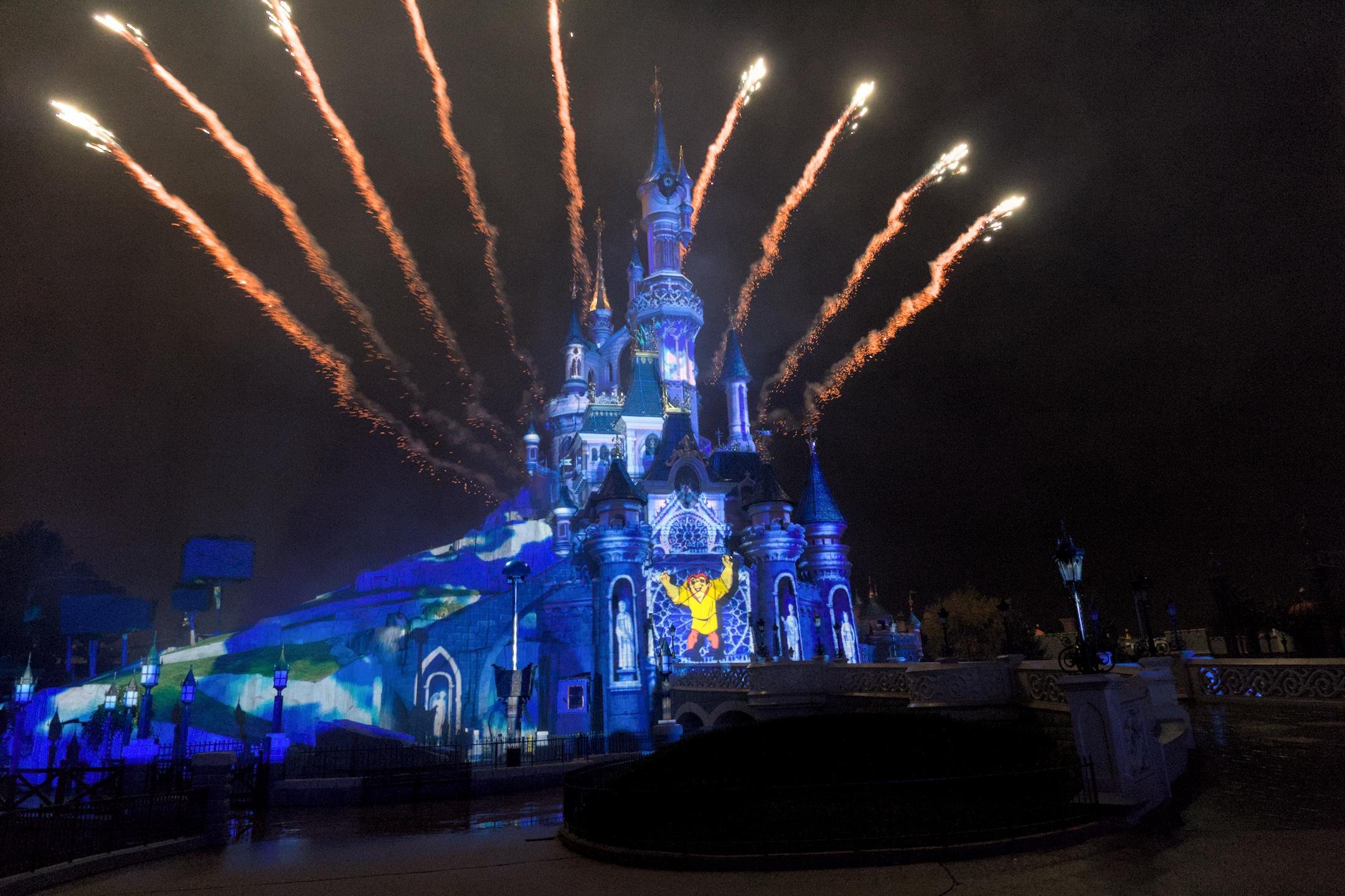 Le château de Disney Paris de nuit, éclairé en bleu, avec 9 traits oranges de feu d’artifice formant un éventail derrière. Le bossu de Notre-Dame est projeté au centre du château