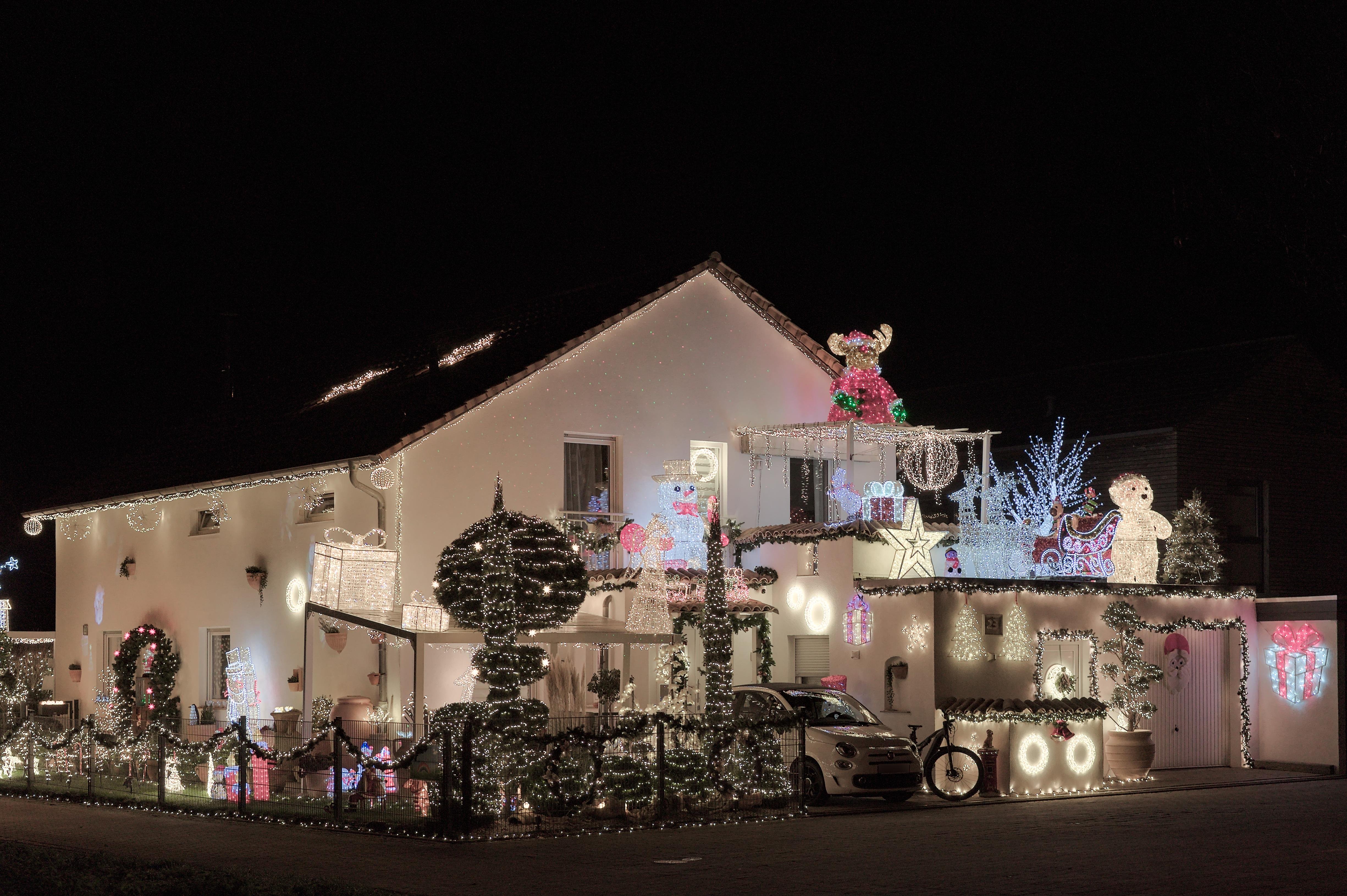 Two-story house at night decorated with Christmas lights, including illuminated wreaths, garlands, light-wrapped trees, and rooftop figures such as a reindeer and snowman; a car is parked in the driveway behind a low fence. ### 
Zweistöckiges Haus nachts, geschmückt mit Weihnachtslichtern, darunter beleuchtete Kränze, Girlanden, mit Licht umwickelte Bäume und Dächerfiguren wie ein Rentier und ein Schneemann; Ein Auto steht in der Einfahrt hinter einem niedrigen Zaun. 