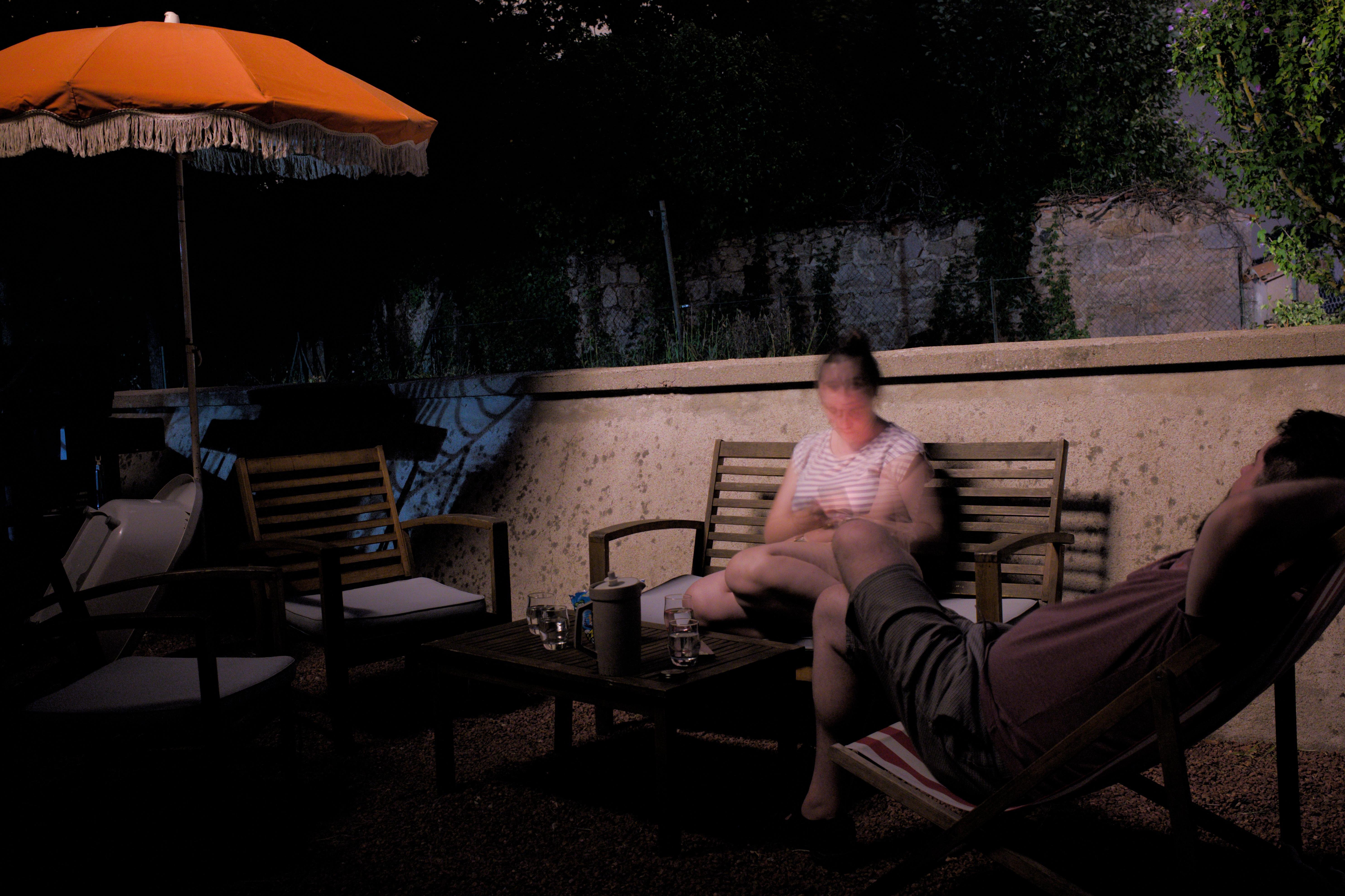 The image shows a nighttime scene of two people sitting on wooden chairs on a patio. There is a table between them with glasses and what looks like a pitcher. An orange umbrella provides some coverage, and there are additional chairs around. The background has a wall with some foliage. The lighting creates a relaxed and dim ambiance. One of the people's faces is blurred from motion.