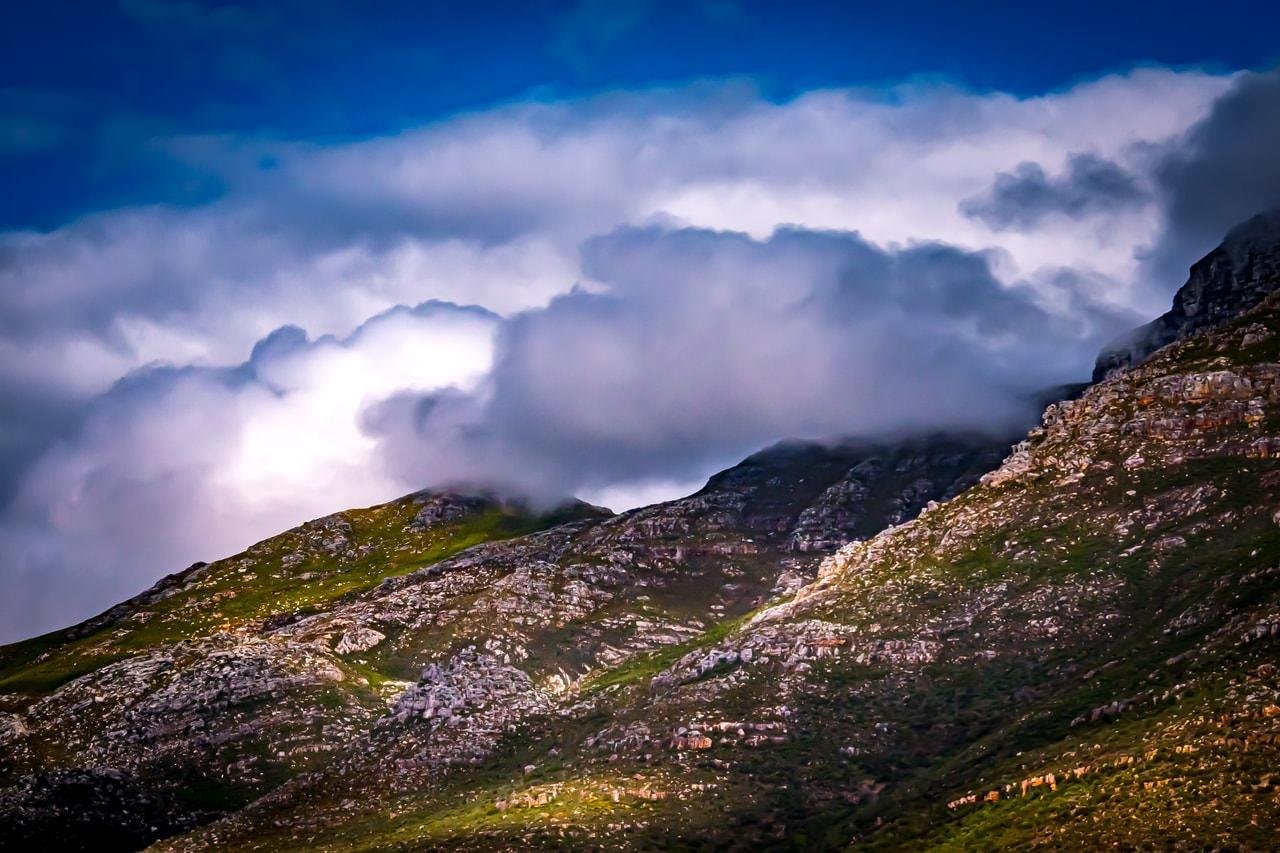 This image shows a landscape of rugged mountains partially covered with green vegetation. Thick clouds hover above the peaks, creating a dramatic and atmospheric scene against a blue sky.