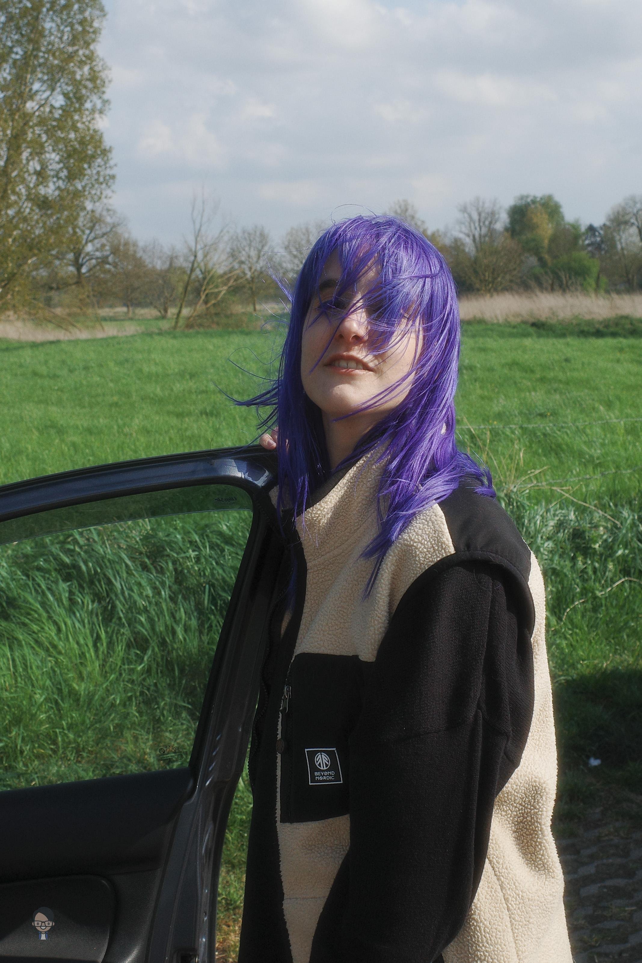 The image shows a person with purple hair standing next to an open car door in a grassy outdoor setting. They are wearing a black and beige jacket. The background includes trees and a cloudy sky.