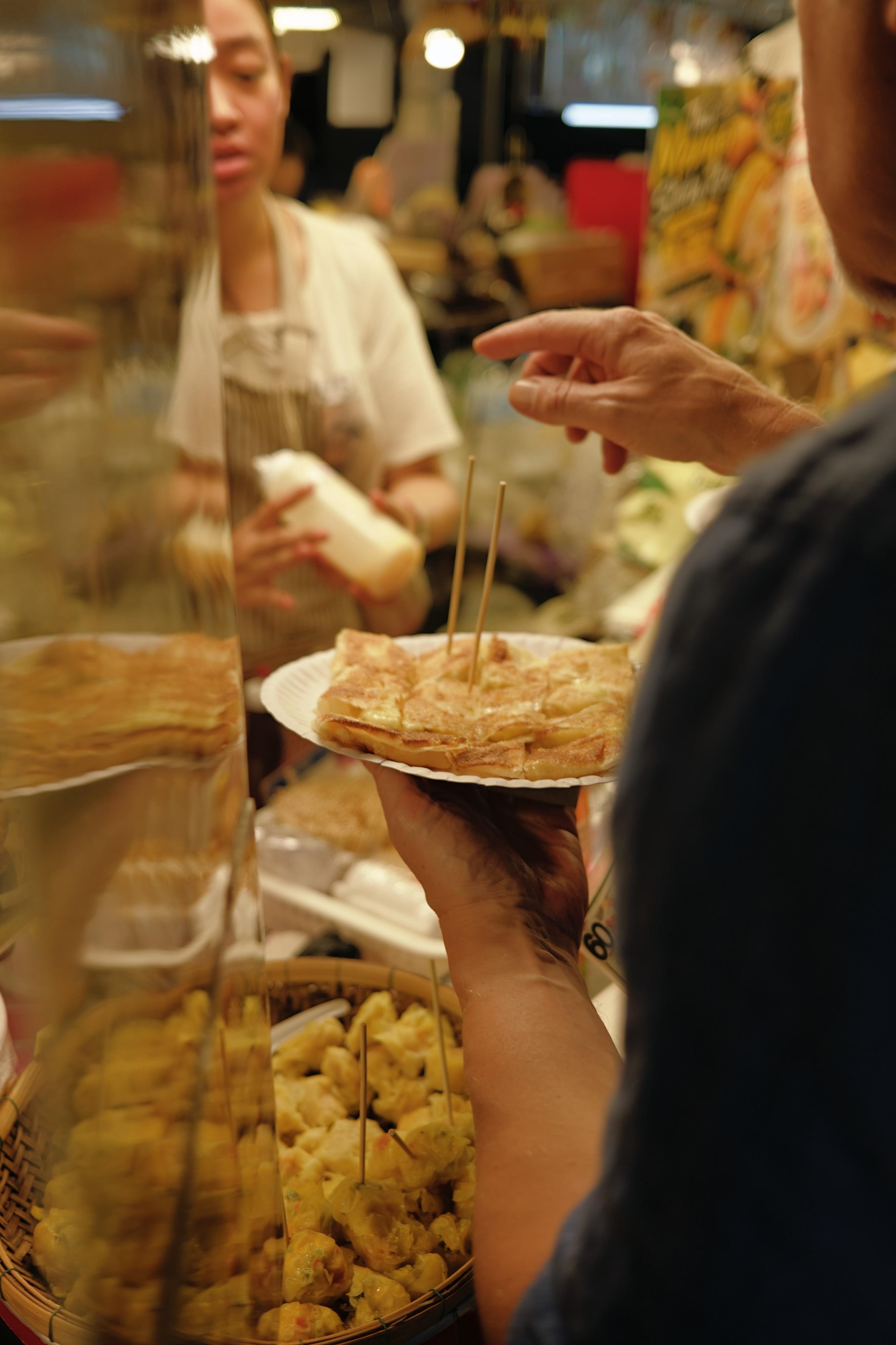 An over-the-shoulder shot of a man holding a plate of crêpes just handed to them by a street vendor. Their other hand is pointing as if they are trying to get something else.