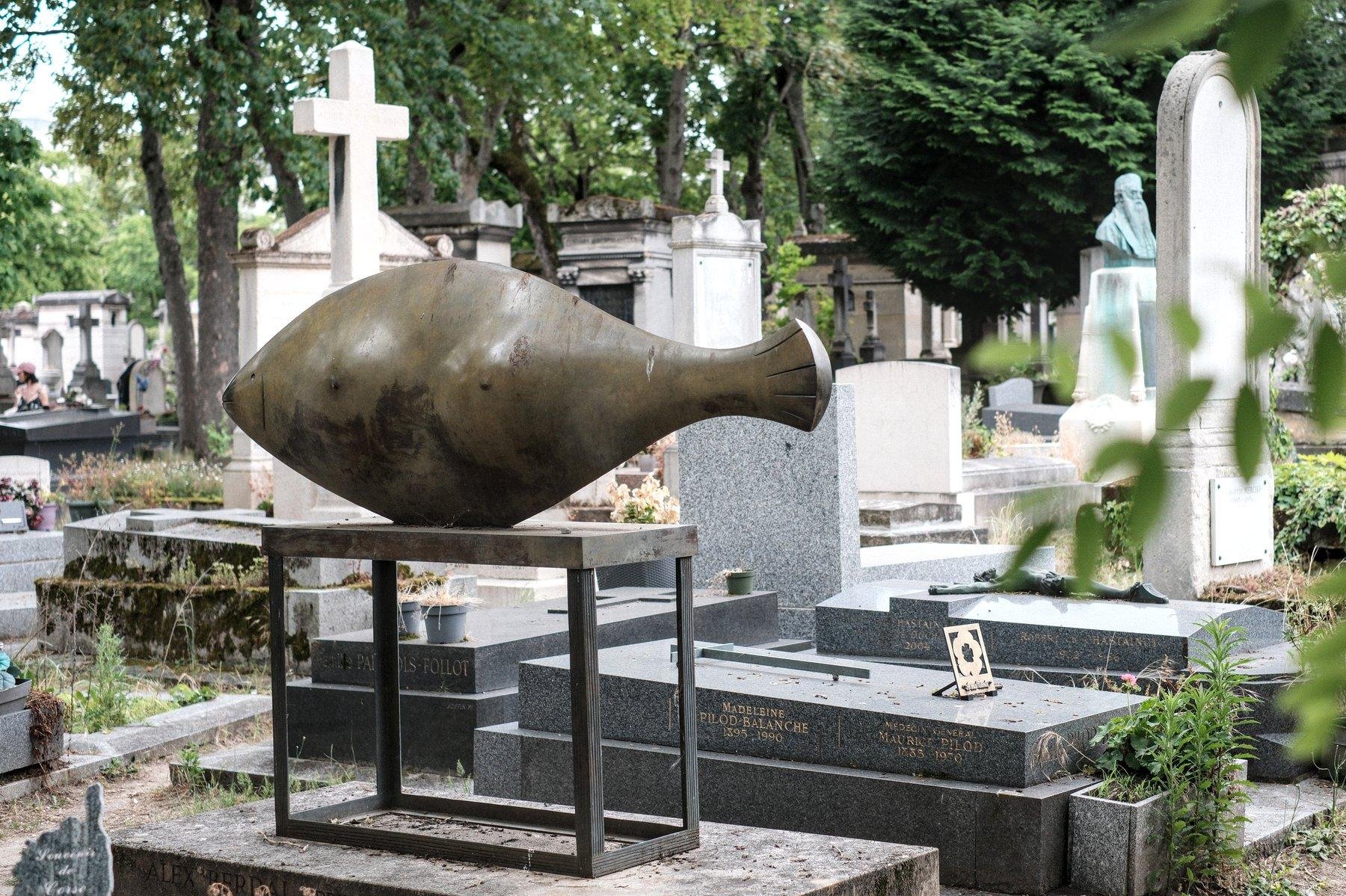 Photo horizontale. Au milieu d’autres tombes, l’une d’entre elle sort du lot avec une décoration en forme de poisson stylisé en métal (bronze ?) posé sur un socle en métal, le tout sur la tombe