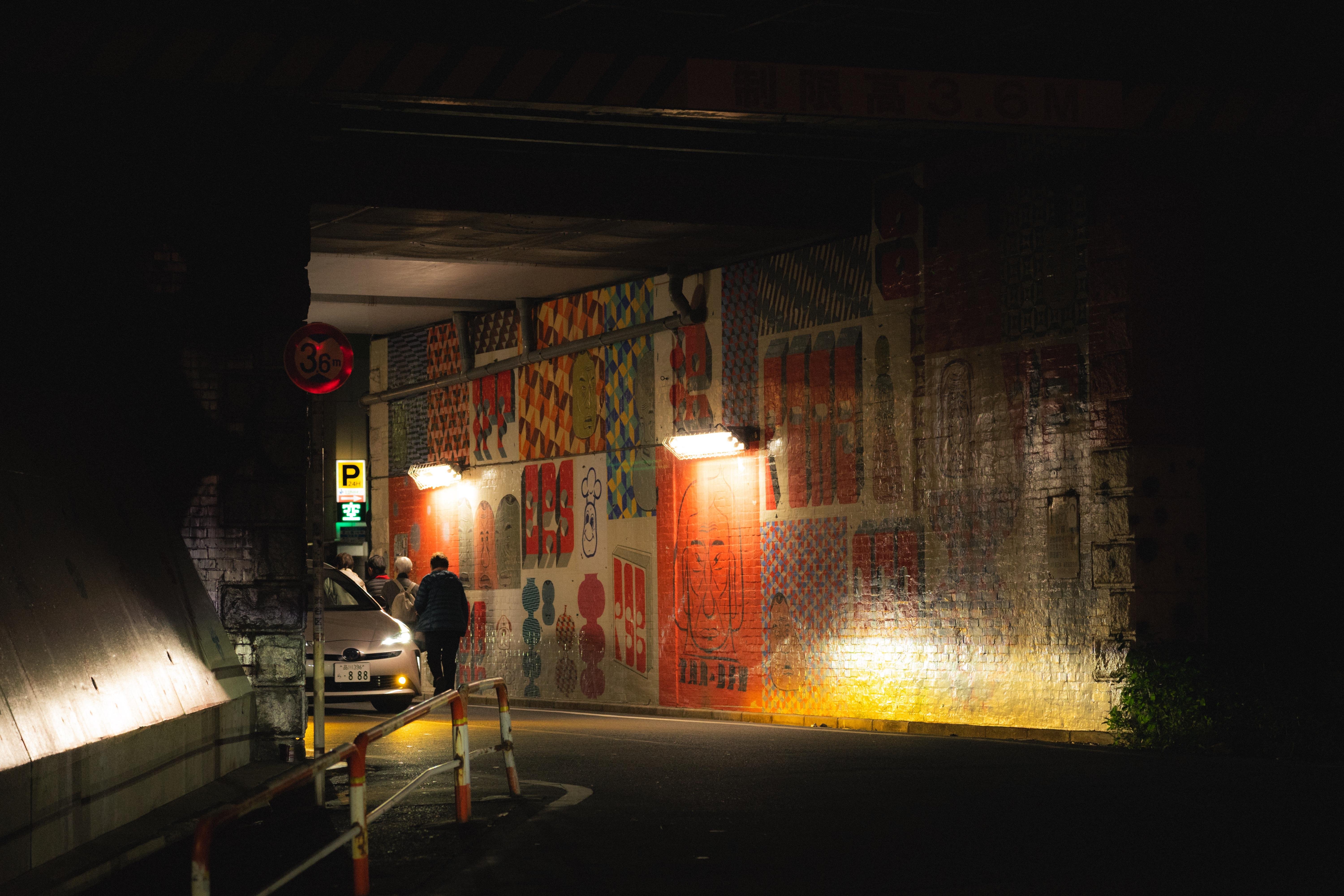 Street underpass at night with brightly lit, colorful mural-covered walls featuring geometric and abstract patterns. Two people walking near a parked car under illuminated street lamps with overhead clearance sign reading 3.6 meters. Dark surroundings contrast the vibrant artwork.