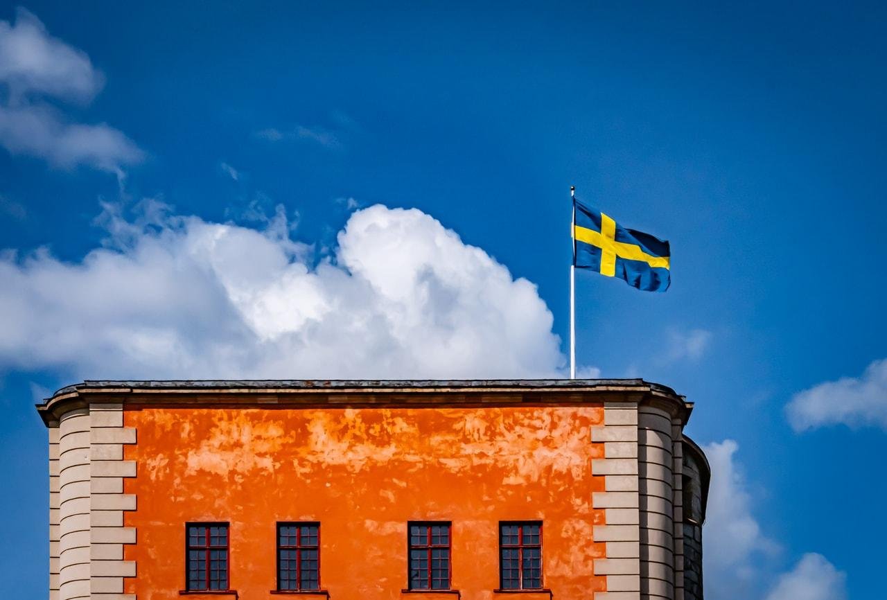 Orange building with a flat roof and beige stone accents, topped with a Swedish flag waving against a blue sky with fluffy white clouds. Four rectangular windows are visible on the facade.