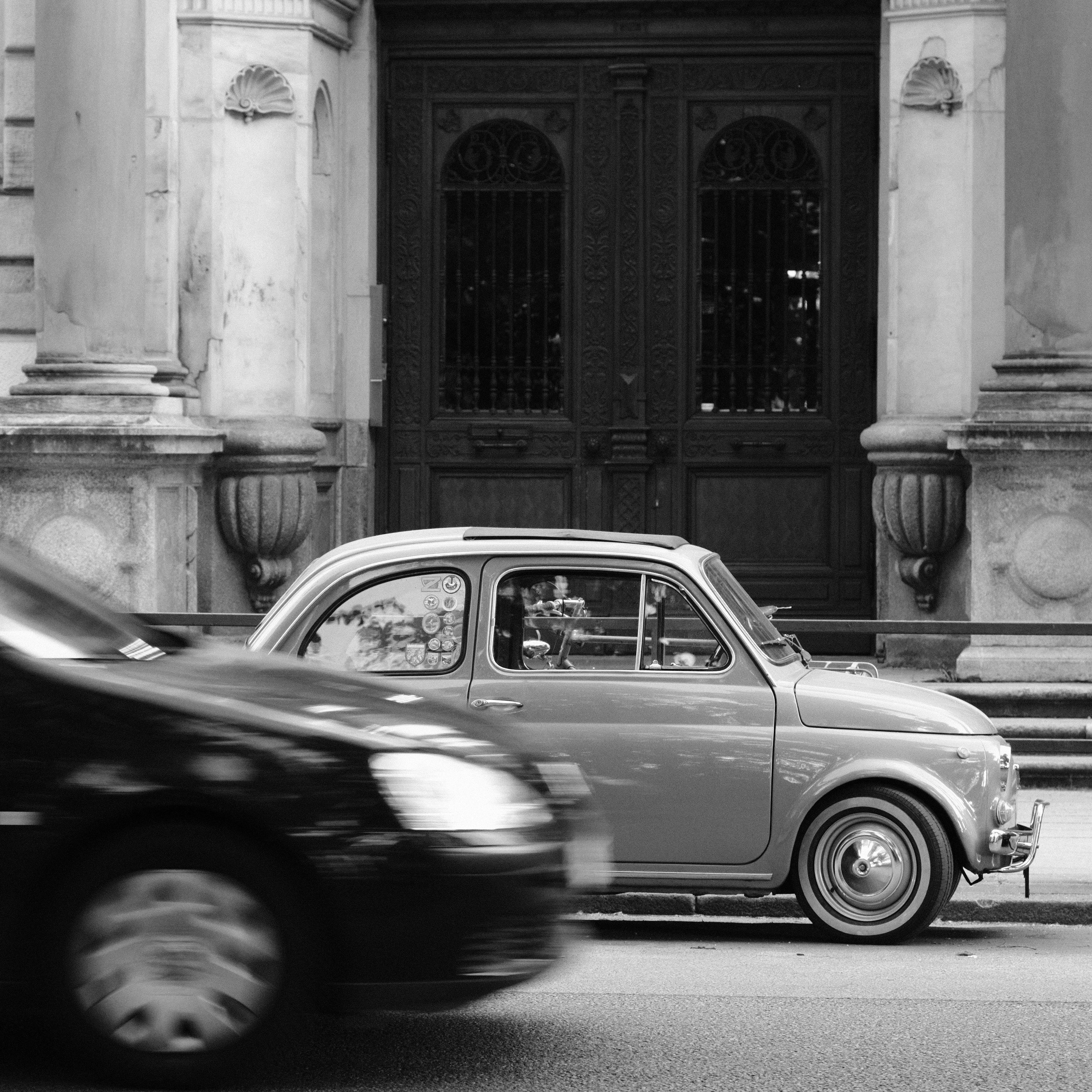 Monochrome / black and white image. A small vintage is car parked in front of a building with marble columns flanking a large wooden entrance door. The photo is taken from the opposite side of the road. A modern dark car is driving on the road between, partly covering the voltage car, adding depth to the scene. Due to the speed the modern car is motion-blurred, contrasting the sharp and focused vintage car.