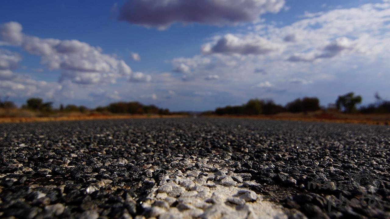 The image shows a low-angle view of an asphalt road under a partly cloudy sky. The foreground focuses on the texture of the road, while the background features trees and a horizon.