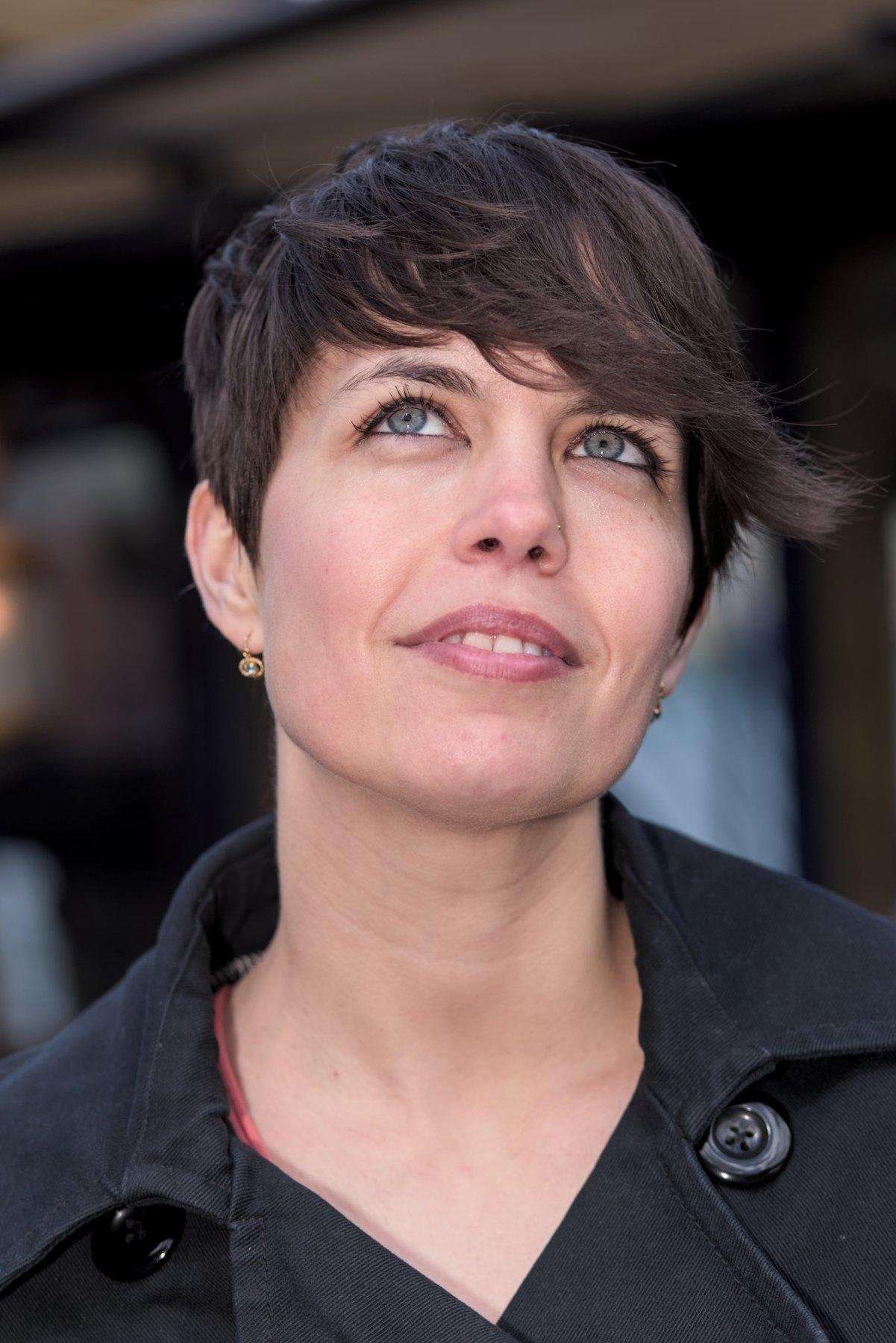 Portrait d’une jeune femme aux cheveux bruns et courts, avec un manteau noir, regardant devant elle et en l’air