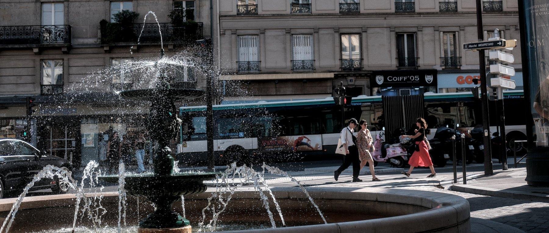 Photo horizontale. Dans la rue, à gauche et au 1er plan, une fontaine allumée avec ses jets figés, à droite au second pan, des gens qui traversent la rue de gauche à droite, croisant une femme avec une longue jupe rouge qui va en sens opposé, en arrière plan, un double bus et des immeubles parisiens typiques