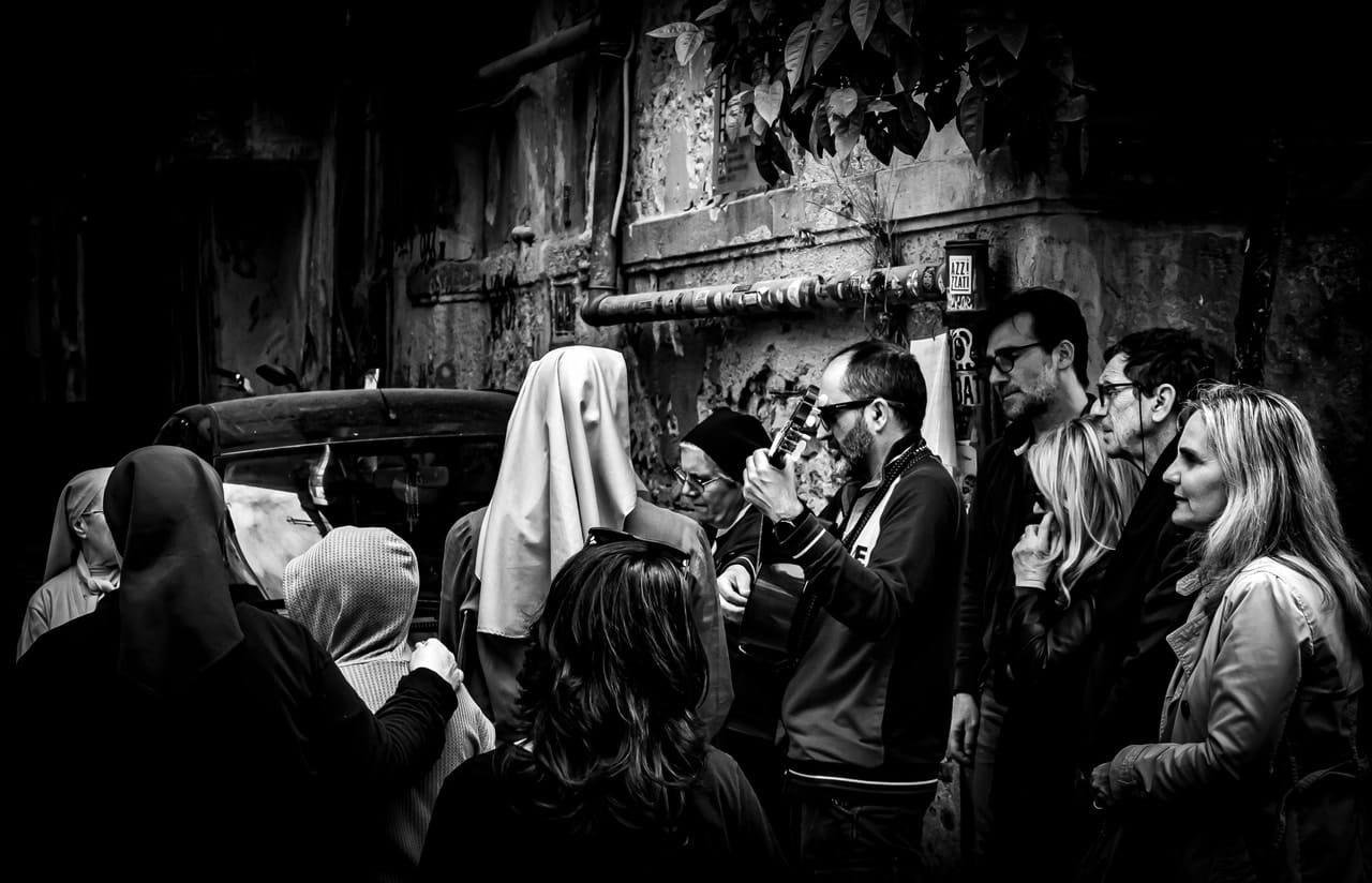 Black-and-white street scene with a small crowd gathered in a narrow alley beside a weathered wall and parked car; a man at center-right plays an acoustic guitar while several people, including women wearing headscarves and a veiled figure, watch from close range.