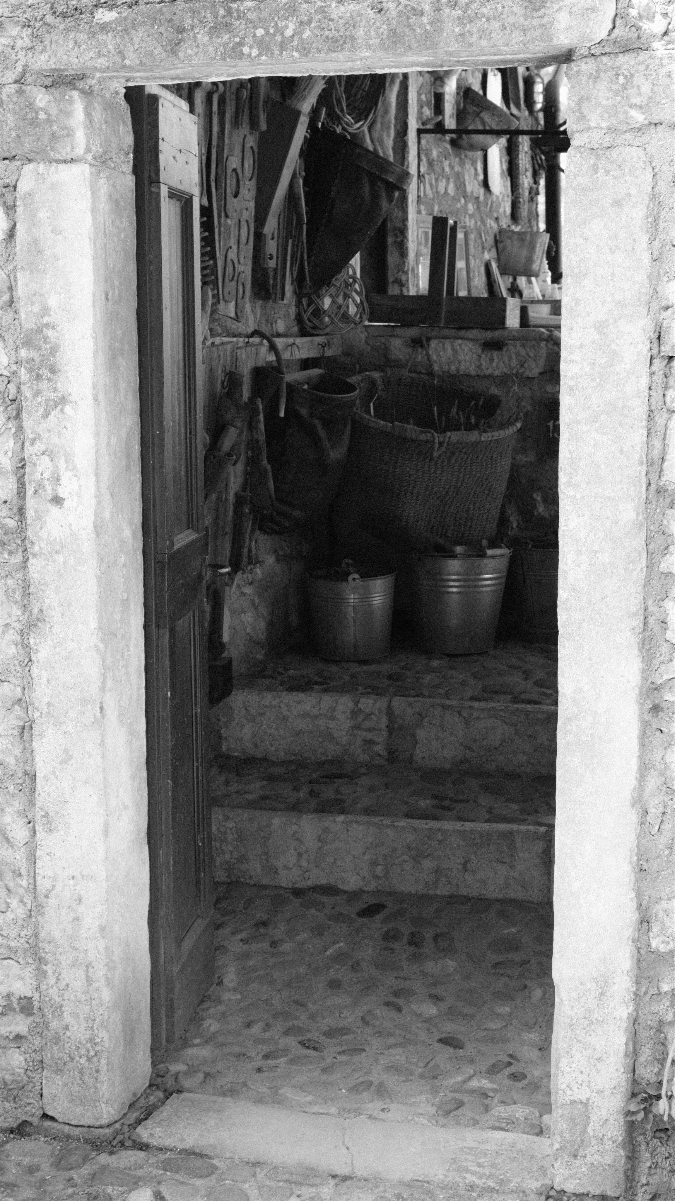 A view through an open wooden door, set into an old stone wall. Behind the door a few steps lead up to a wall hung with rusty tools, and baskets full with nails, metal parts and smaller tools. All in monochrome black and white.