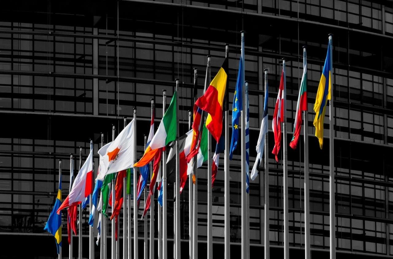 Multiple national flags on poles arranged in a row, set against the backdrop of a glass building facade. Flags include various colors and patterns, prominently featuring reds, blues, yellows, and greens. The architecture in the background reflects modern design with numerous windows.