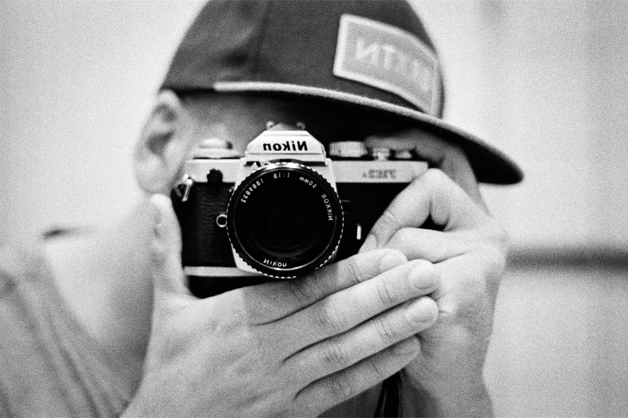 Person wearing a hat with the brim lowered, peering through a vintage Nikon film camera, framed in a black and white photograph. Hands are visible holding the camera, focusing on capturing an image. The classic bathroom mirror selfie!
