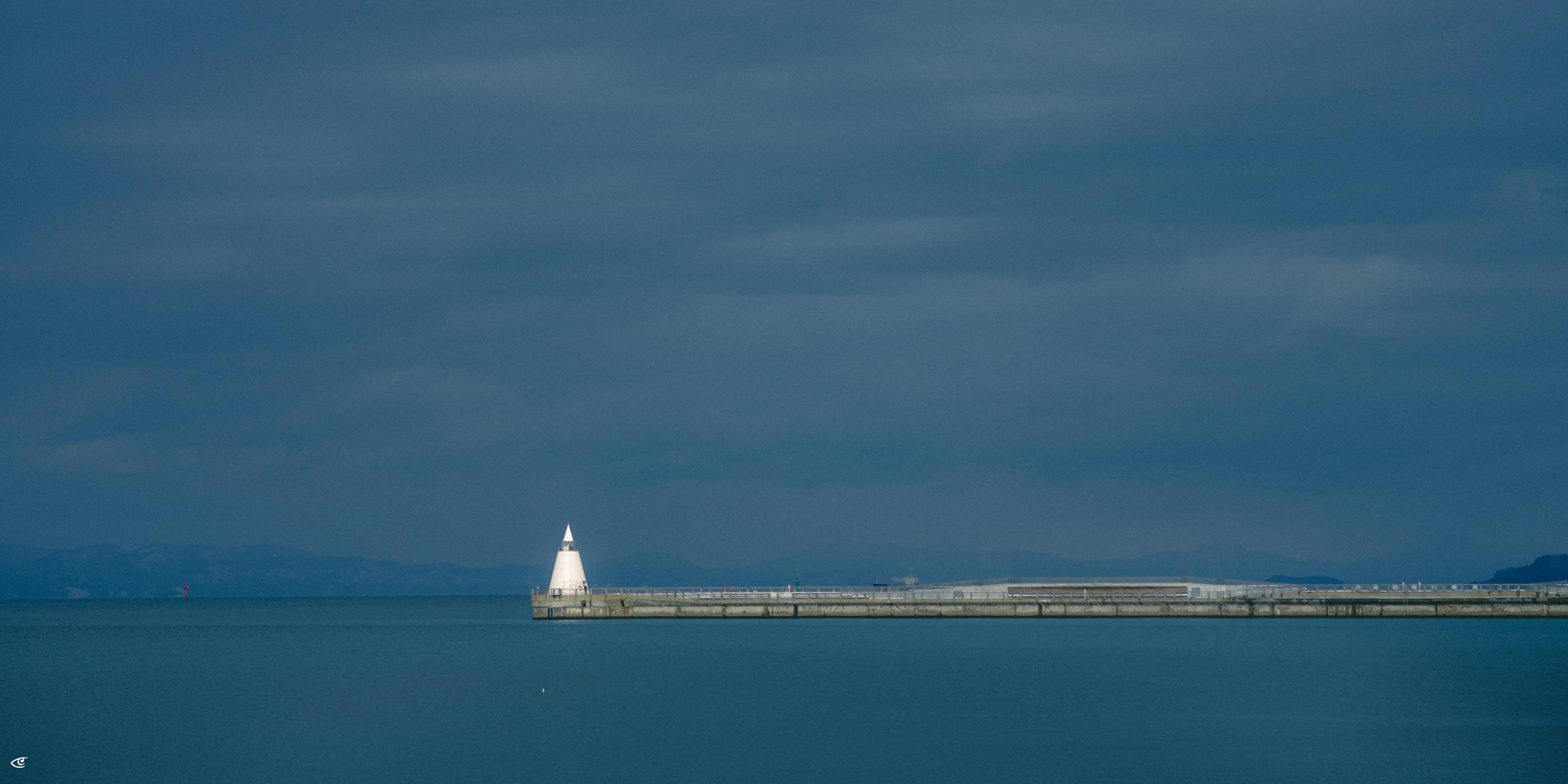 White lighthouse at the end of a long concrete breakwater extending from right to left across calm blue water, with dark cloudy sky and faint distant mountains on the horizon.