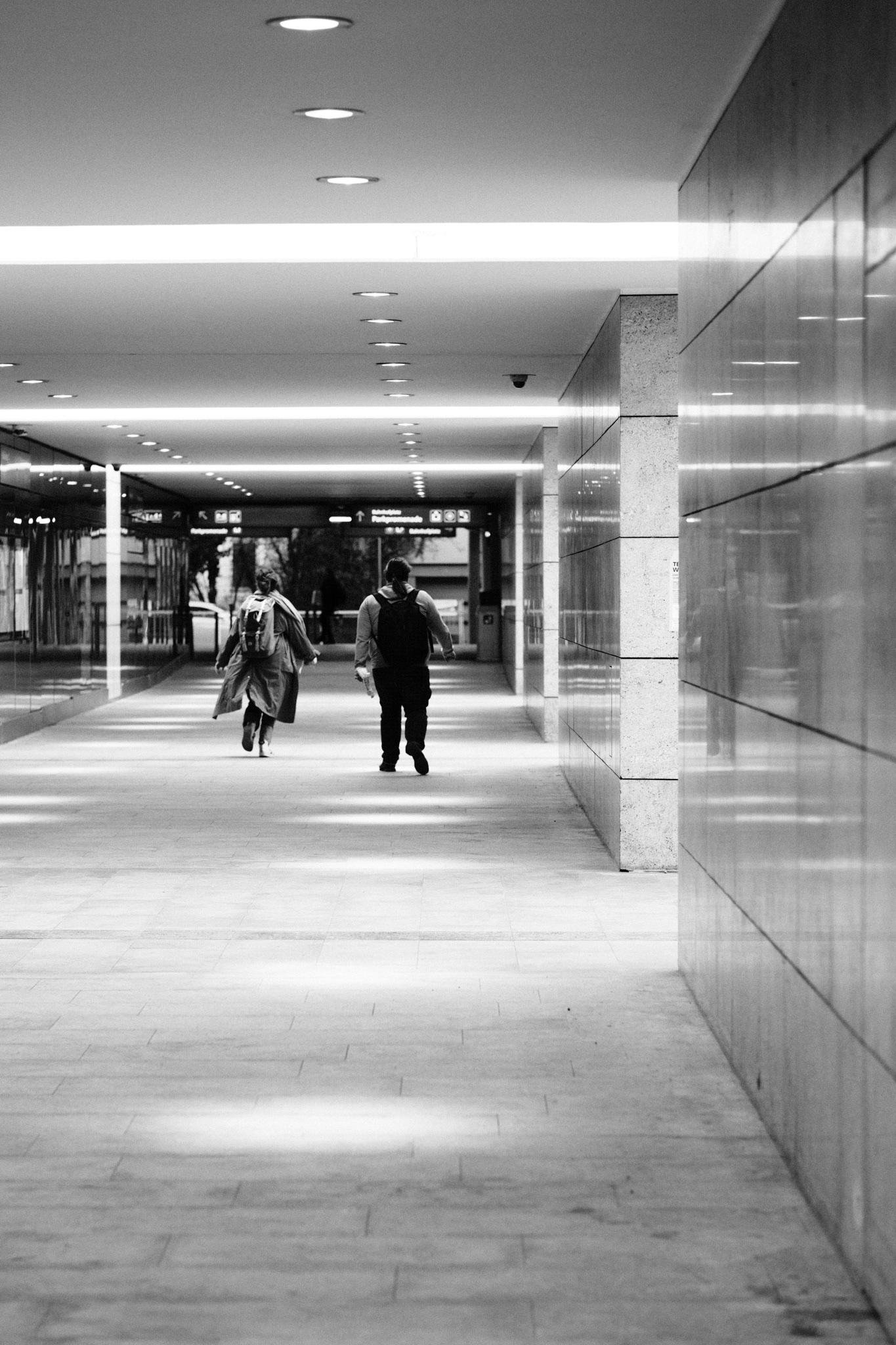 Schwarz-weiß Foto  Blick hinunter auf einen langen überdachten Korridor mit Fliesenboden, reflektierenden Wandpaneelen auf der rechten Seite und einer Reihe von Einbaudeckenleuchten; zwei Personen mit Rucksäcken gehen in Richtung entfernter Überkopfschilder.