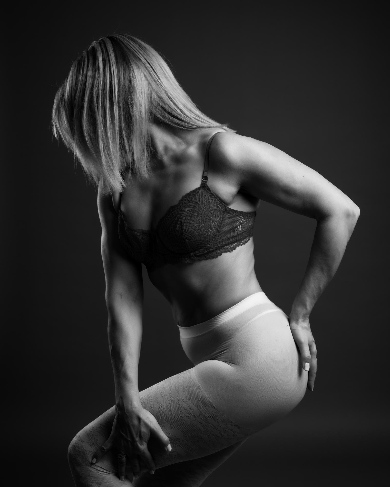 The image is a black-and-white photograph depicting a woman in a side pose. She is wearing a patterned bra and high-waisted tights. The image focuses on her muscular physique and pose.