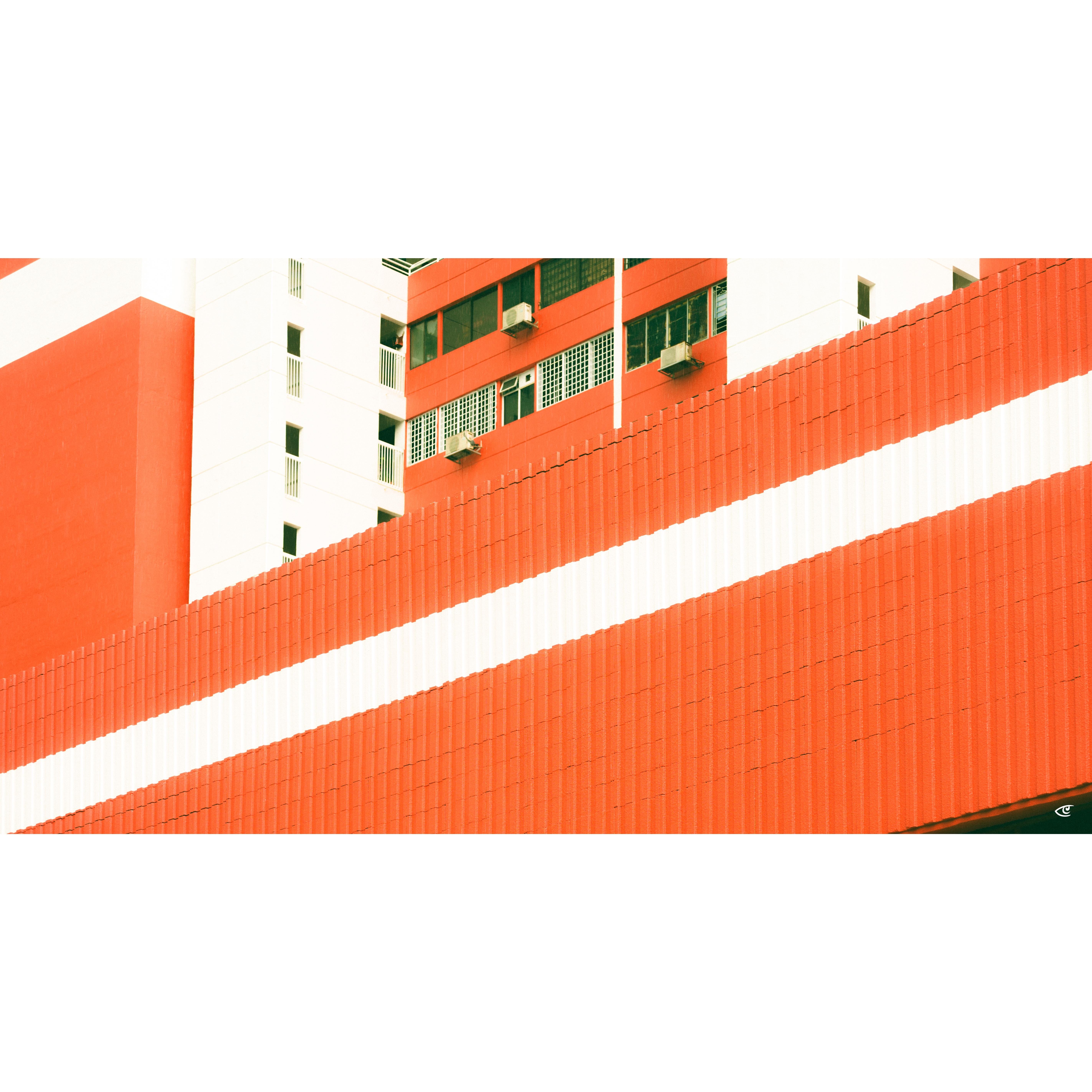 Low-angle view of a red-and-white building facade, with a wide diagonal red wall in the foreground and upper floors showing windows, grilles, and several air-conditioning units.
