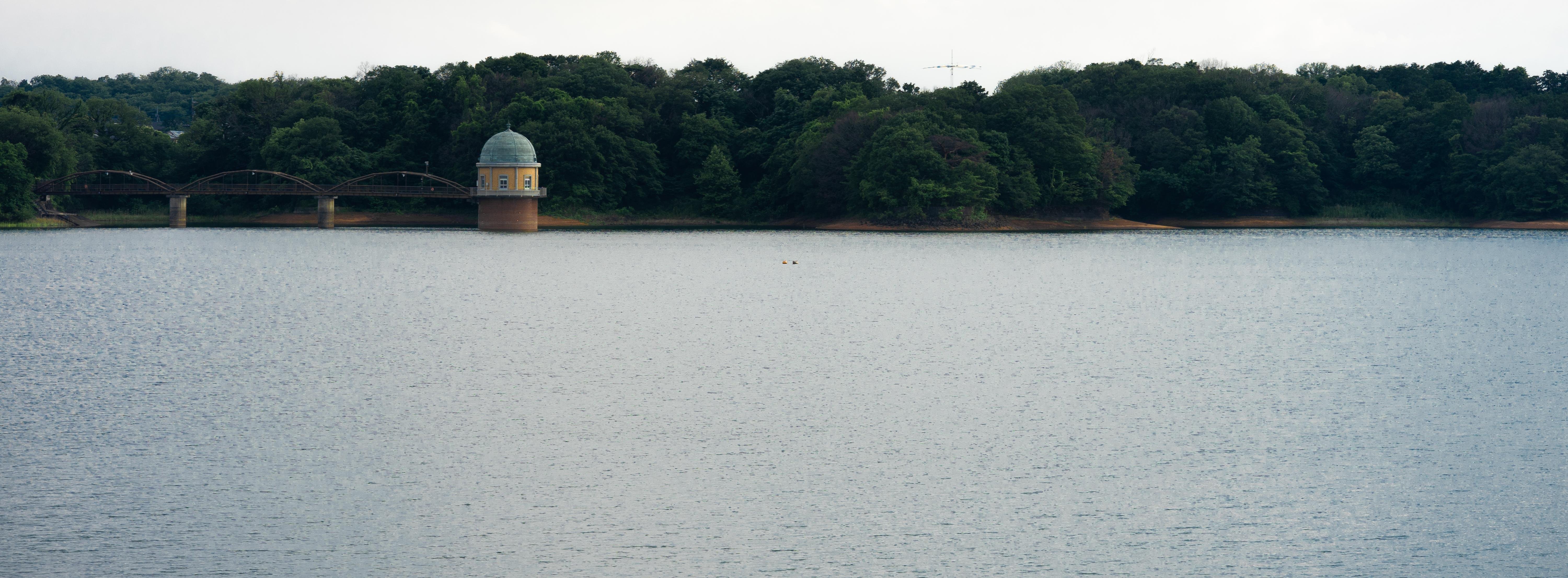 Calm body of water with a small, domed building on stilts near the shoreline, surrounded by dense, green trees. A bridge with multiple arches connects the building to the forested land. Overcast sky above, with the distant outline of hills in the background.