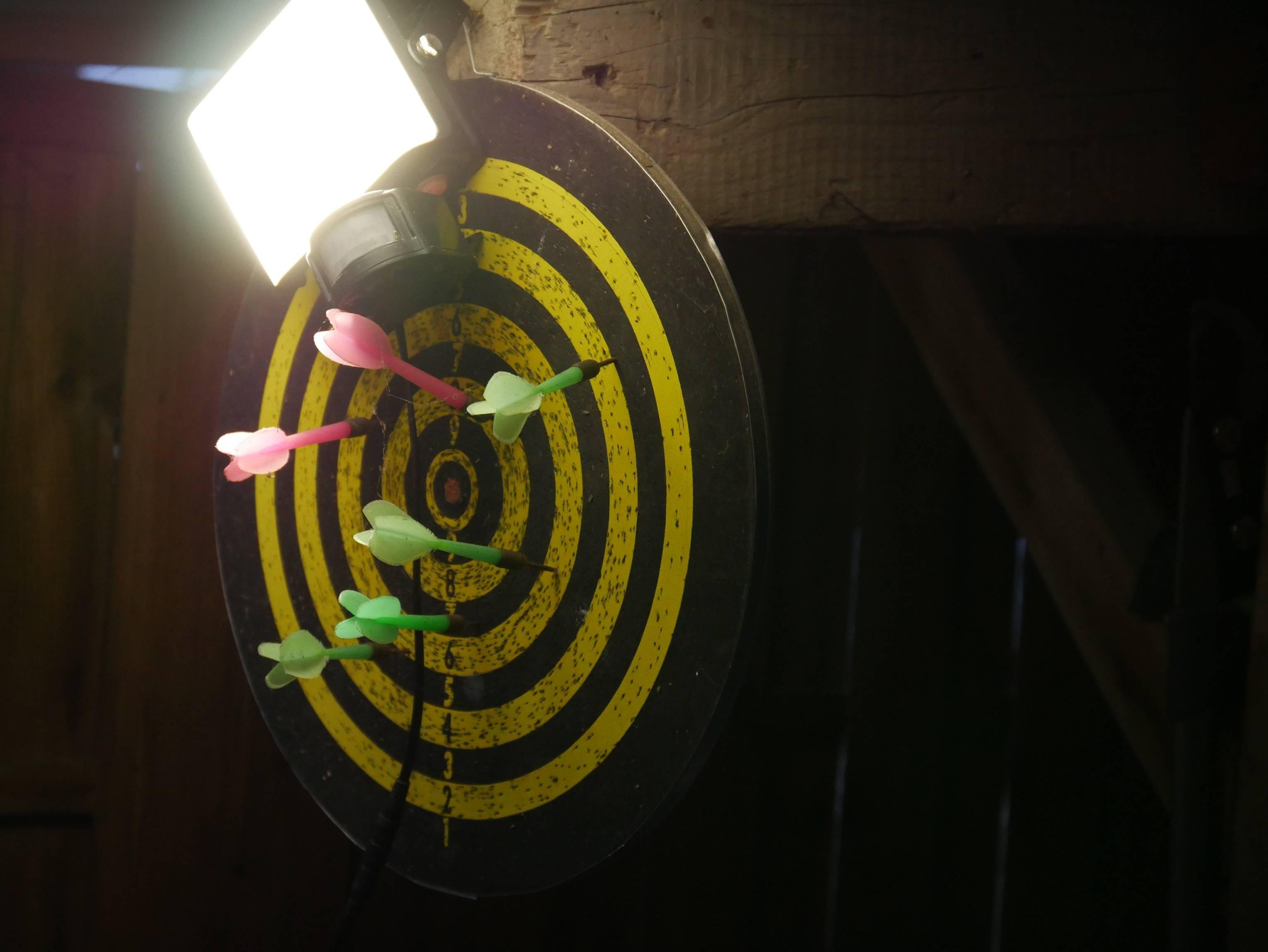 The image shows a dartboard with several brightly colored darts stuck in it. The board has concentric yellow and black circles, typical of target designs. There is a light source shining brightly above the dartboard.