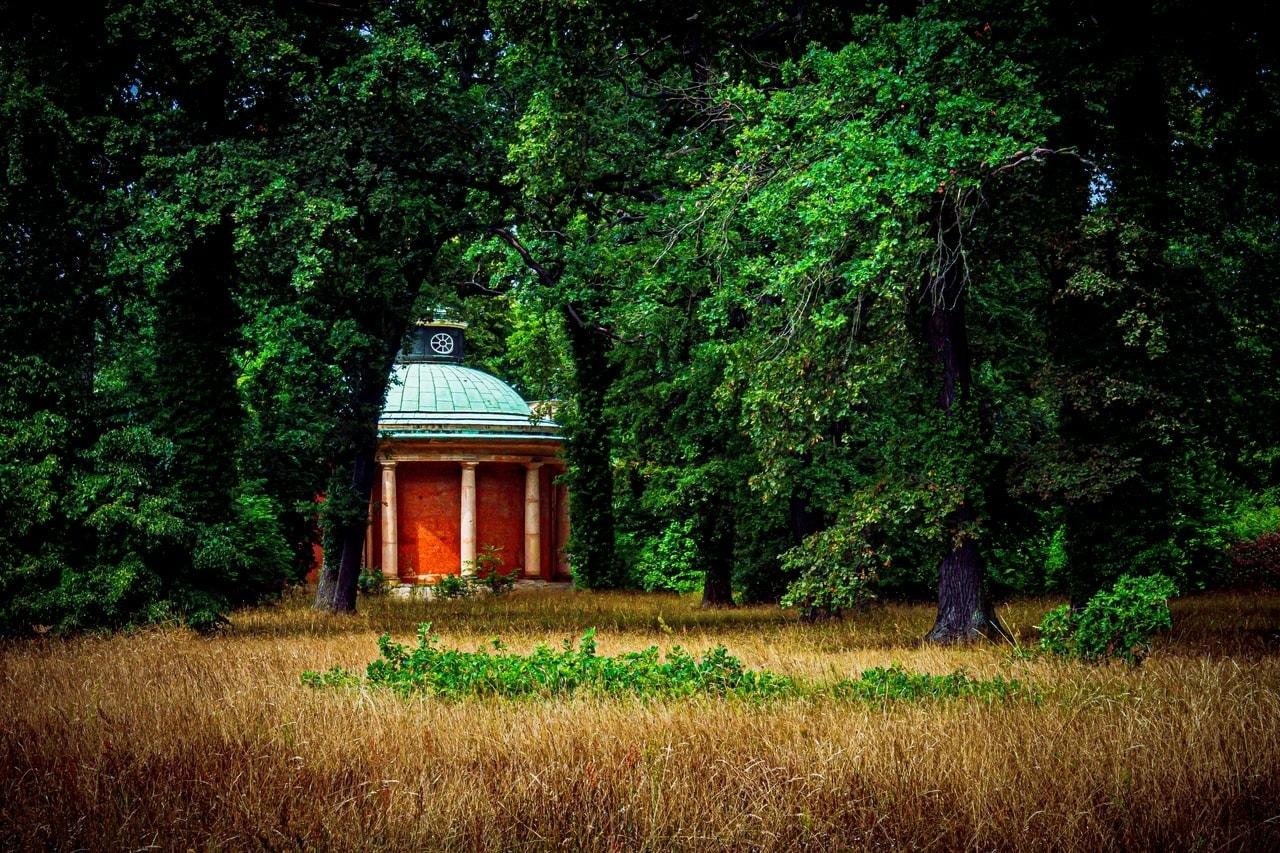 Small neoclassical pavilion with a pale green domed roof and four columns, partially hidden among dense green trees, with tall dry grass in the foreground.