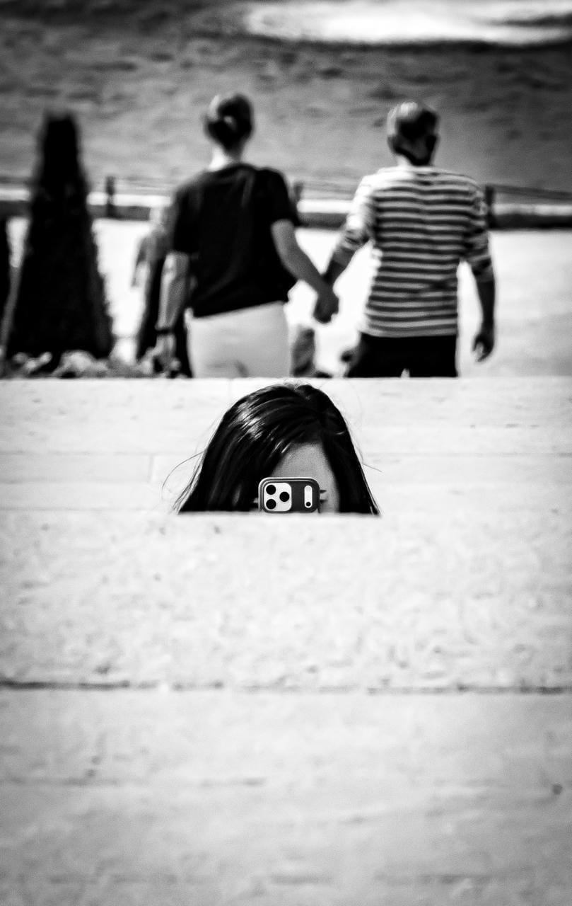 Person with long dark hair peeking over a concrete ledge while holding a smartphone up to their face; in the blurred background, two people walk away holding hands near a waterfront.