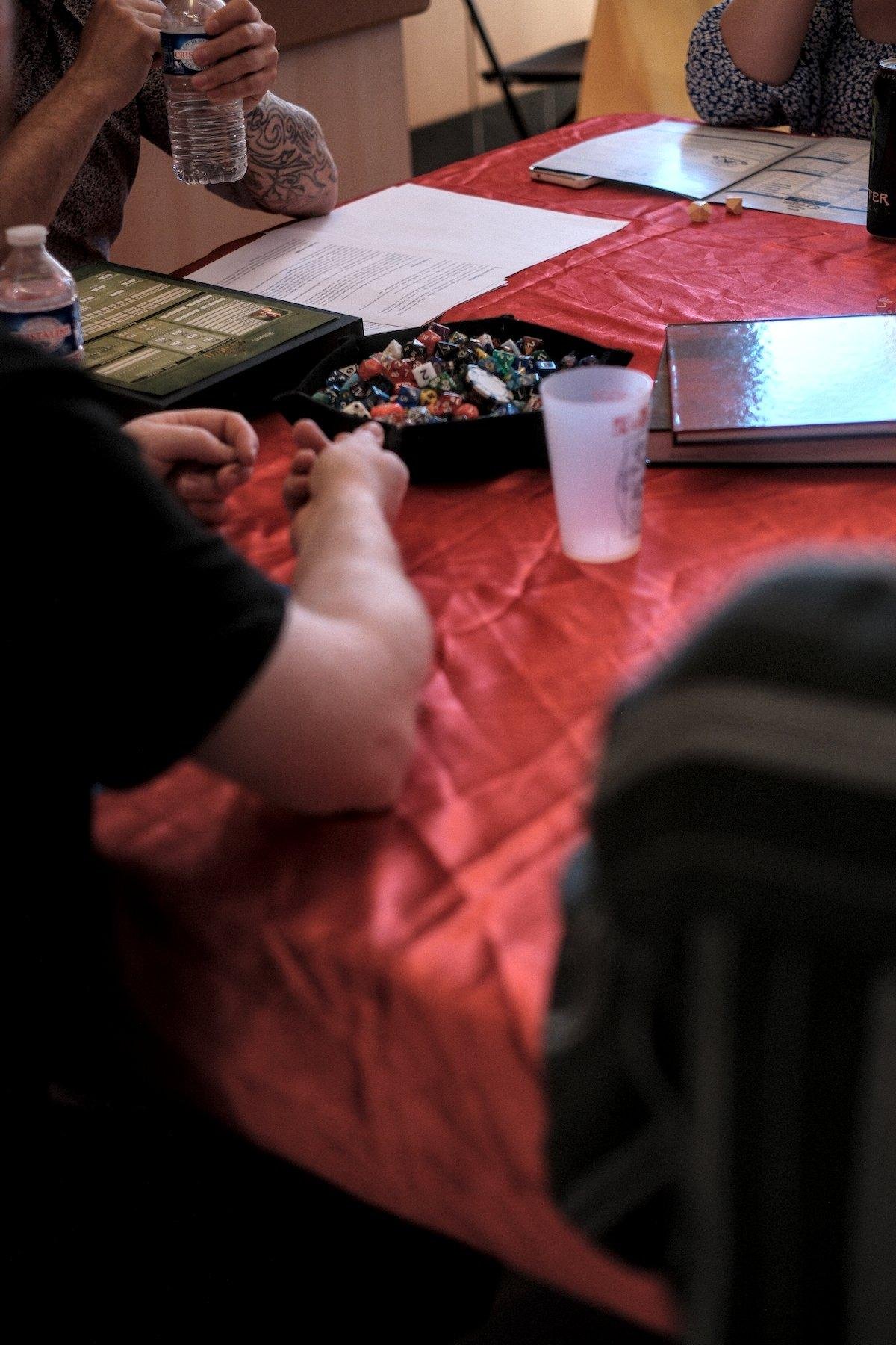 Photo verticale. Une table avec une nappe rouge satiné, avec des gens autour dont on re voit que les épaules, le sujet étant un bac en cuir plein de dé de jeu de rôle