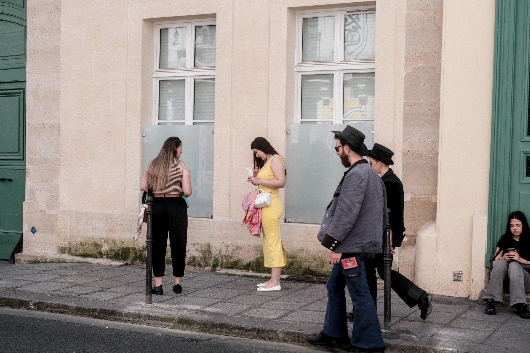 Photo horizontale. Dans la rue, 2 hommes avec chapeau noir, pantalons patte d’éléphant, l’un en denim l’autre noir, une veste grise et l’autre noir. Ils marchent vers la gauche en allant sur la chaussée pour éviter 2 jeunes femmes arrêtées car l’une s’est a priori tâchée avec sa boisson. Sur la droite, une femme assise sur le pas d’une porte verte consulte son téléphone
