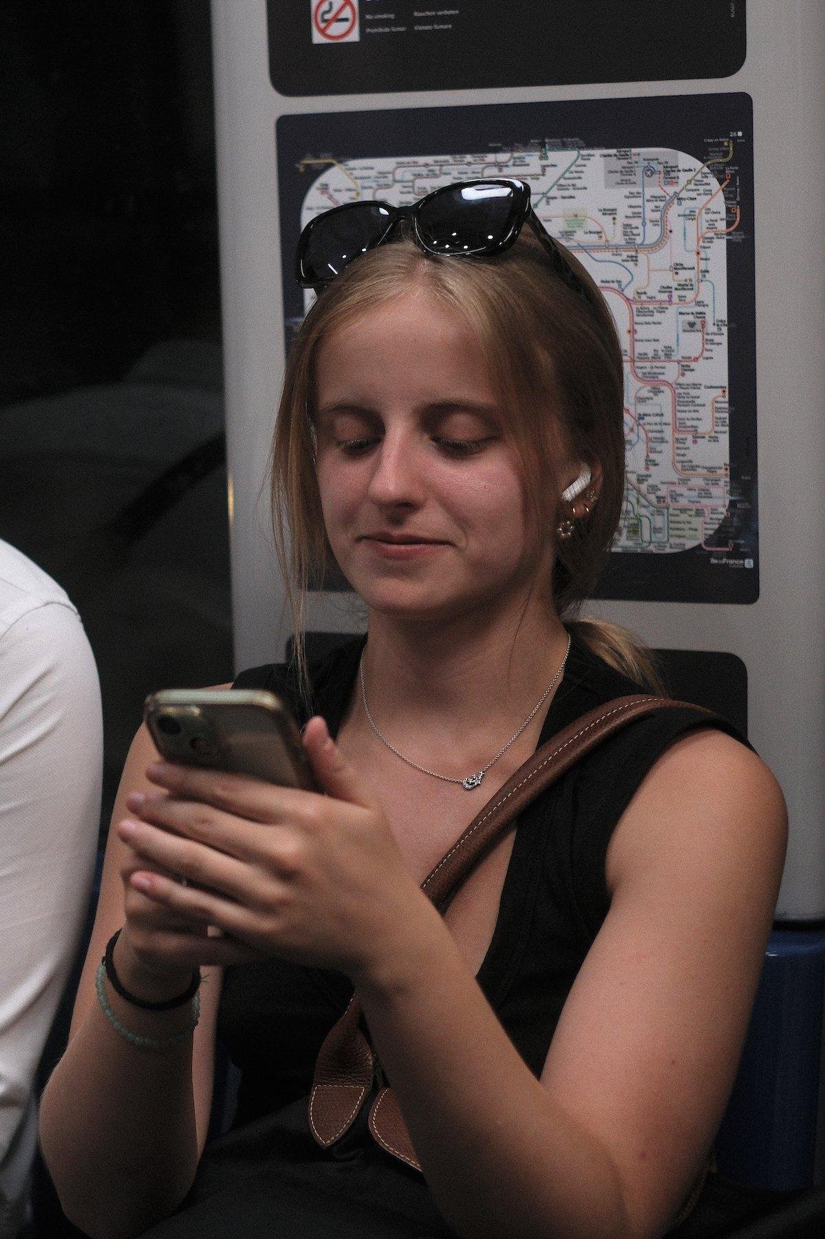 Photo verticale. Une jeune femme châtain, lunettes de soleil posées sur la tête, assise dans le métro, entrain de discuter sur son téléphone avec un petit sourire pincé