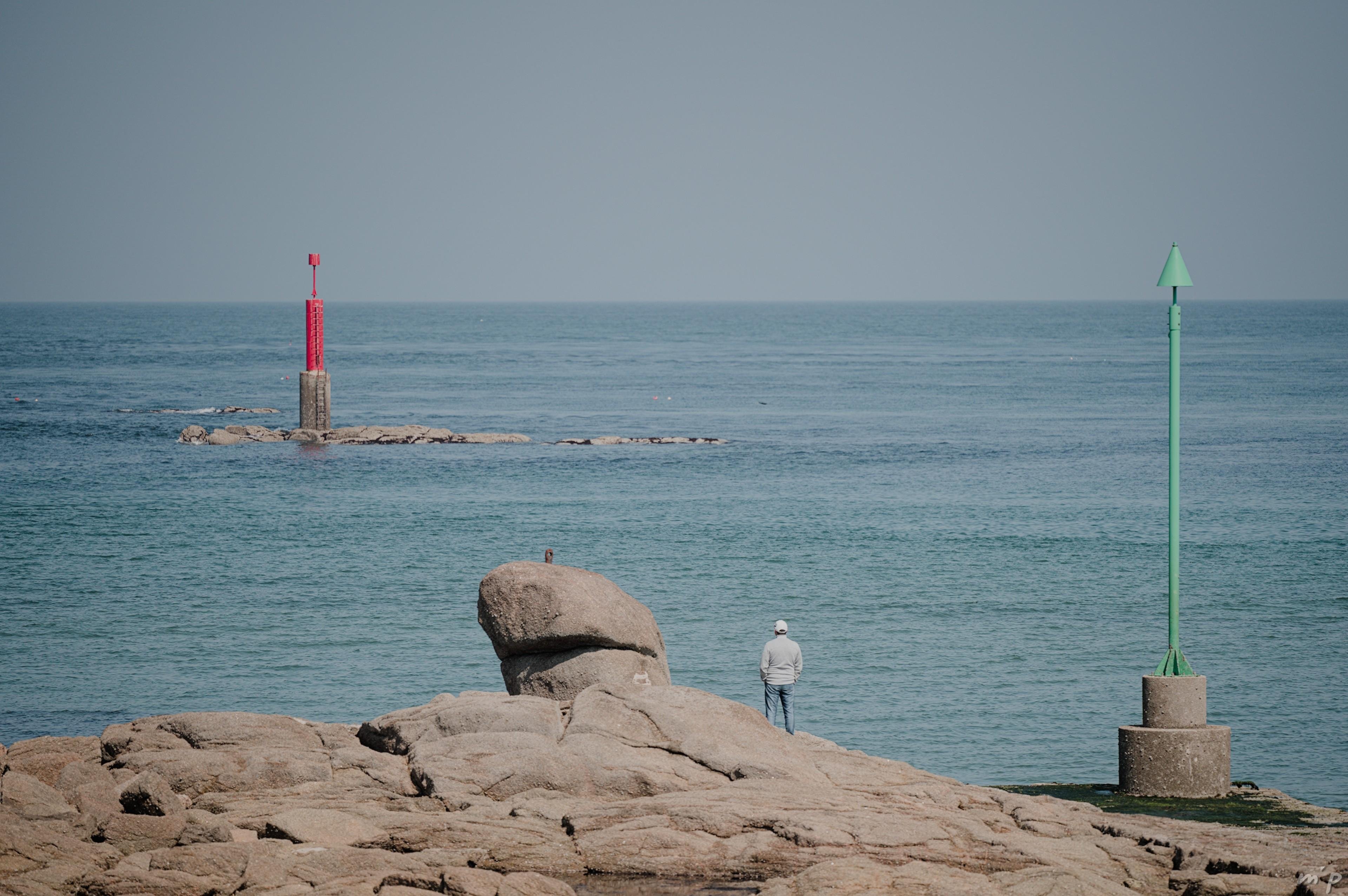 🇬🇧 Horizontal color photo of a motionless man standing from behind, hands in his pockets, on a rocky coastline, facing the calm sea. To the right of the man, on the same rocks a green beacon (cone), to his left on a rocky islet, a red beacon (cylinder).

🇫🇷 Photo couleur horizontale d'un homme immobile debout de dos, les mains dans les poches, sur un littoral de rochers, face à la mer calme. À droite de l'homme, sur les mêmes rochers une balise verte (cône), à sa gauche sur un îlot rocheux, une balise rouge (cylindre).