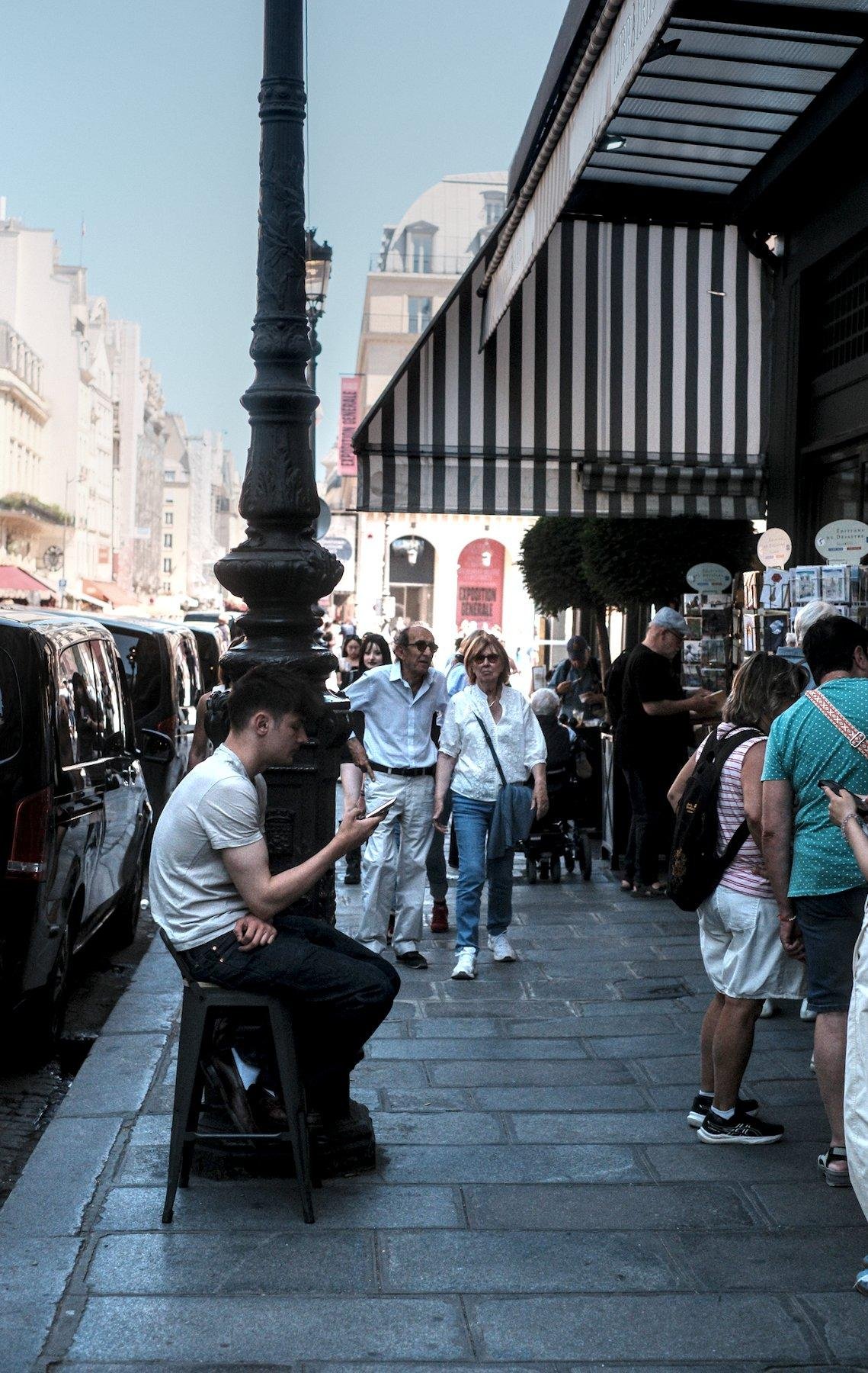 Photo verticale. Sur un trottoir parisien, un jeune homme brun, t-shirt blanc et jean noir, est assis sur un tabouret à côté d’un lampadaire en consultant son téléphone. Des gens passent devant lui sans y prêter attention