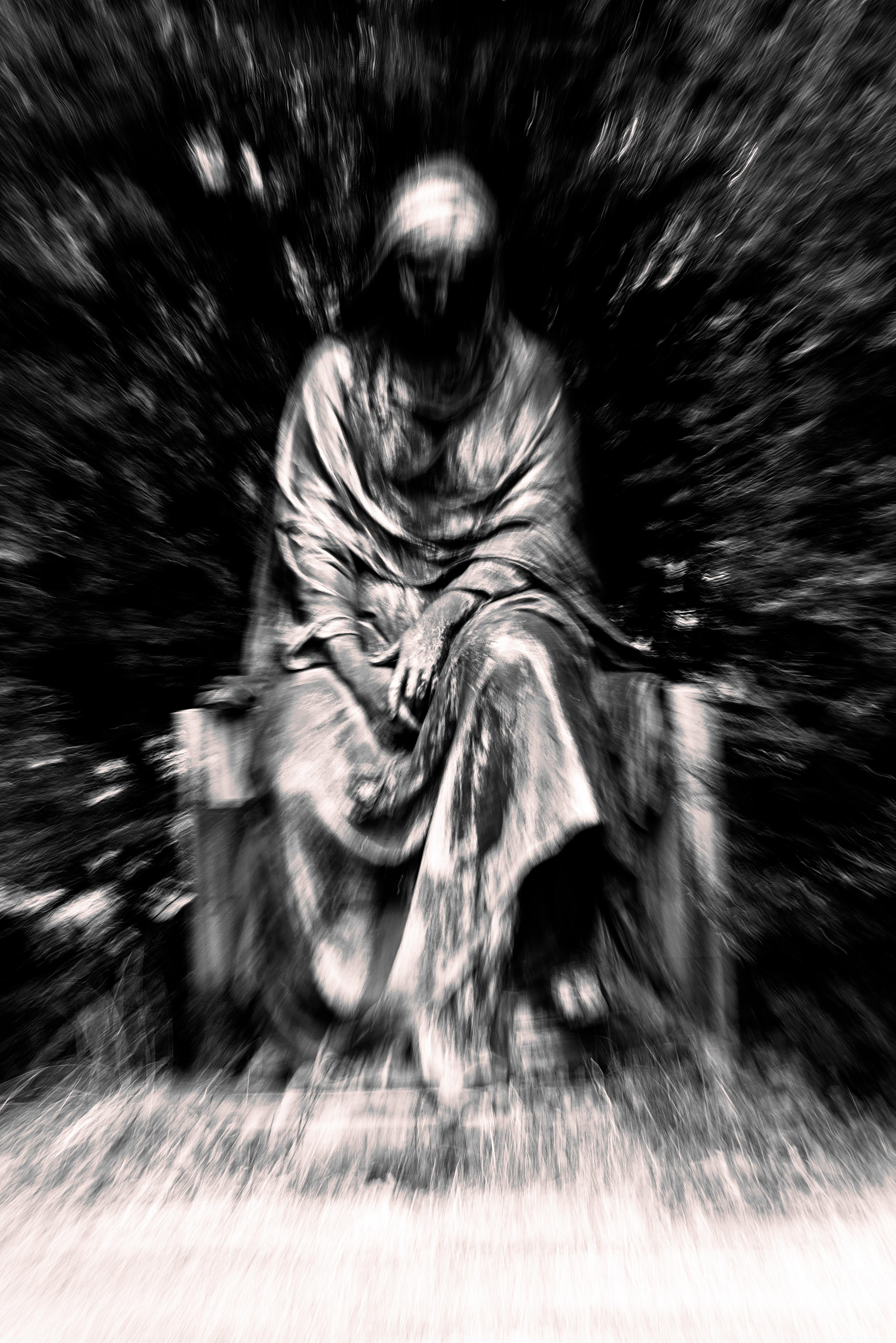 This image depicts a statue of a seated figure draped in cloaked robes. The photo is black and white, with a blurred effect giving it an eerie and mysterious atmosphere. The statue's details are somewhat obscured by the blur, focusing on its contemplative posture.