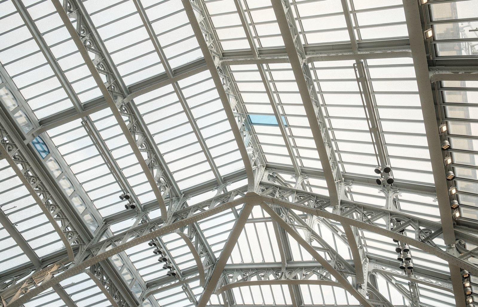 Photo horizontale. Une grande verrière avec ses multiples poutres en acier et support de vitres diffusant la lumière blanche de l’extérieure, sauf une, qui est bleue