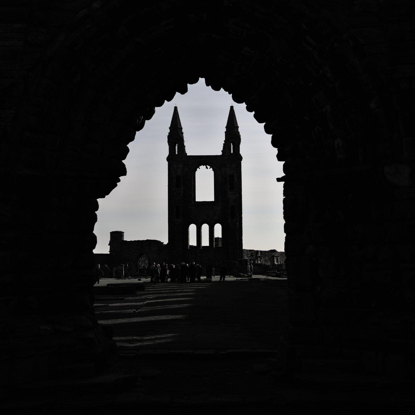 Photo carrée montrant en contre jour les ruines d’une façade de cathédrale, à travers une alcove en pierre avec l’arche caractéristique en pointe de l’architecture gothique