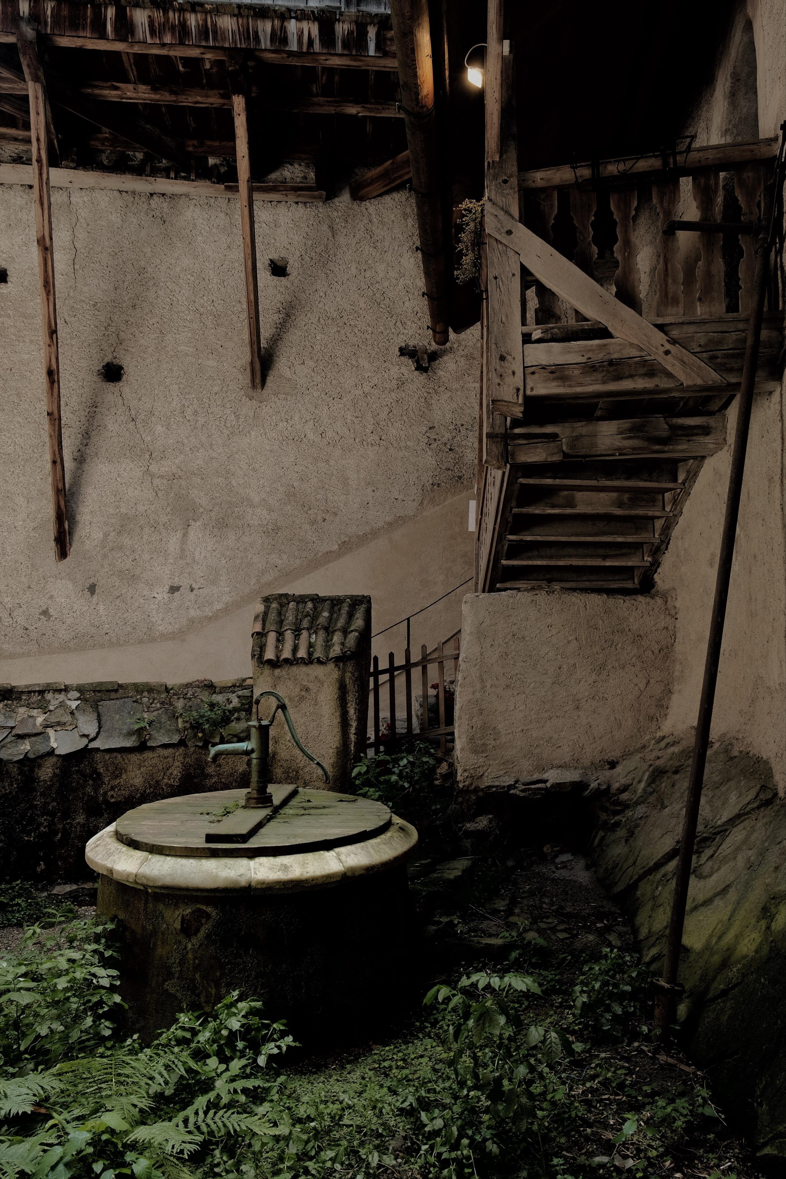 Round stone well with a closed wooden lid and hand pump in the foreground, surrounded by overgrown low plants. Weathered plaster walls in the background and a wooden staircase leading to an upper level in a dim castle courtyard.