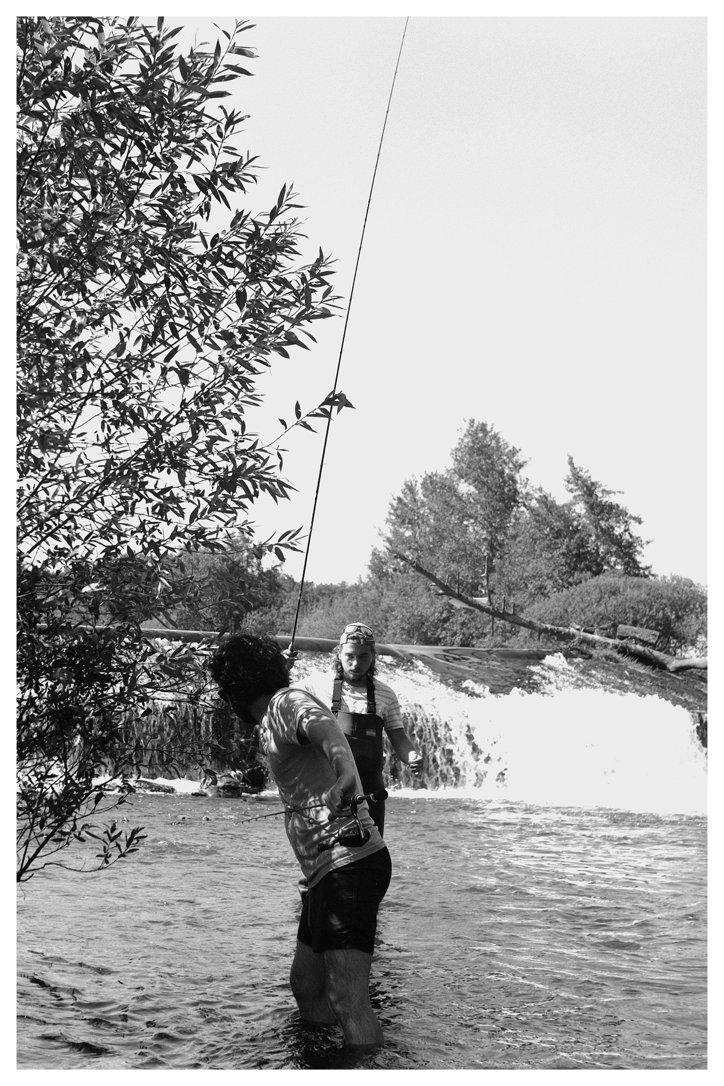 Black-and-white photo of two people standing knee-deep in a river below a small waterfall; one in the foreground holds a fishing rod with the line angled upward, while the other faces the camera. Leafy tree branches fill the left side, with trees and rocks along the riverbank in the background.