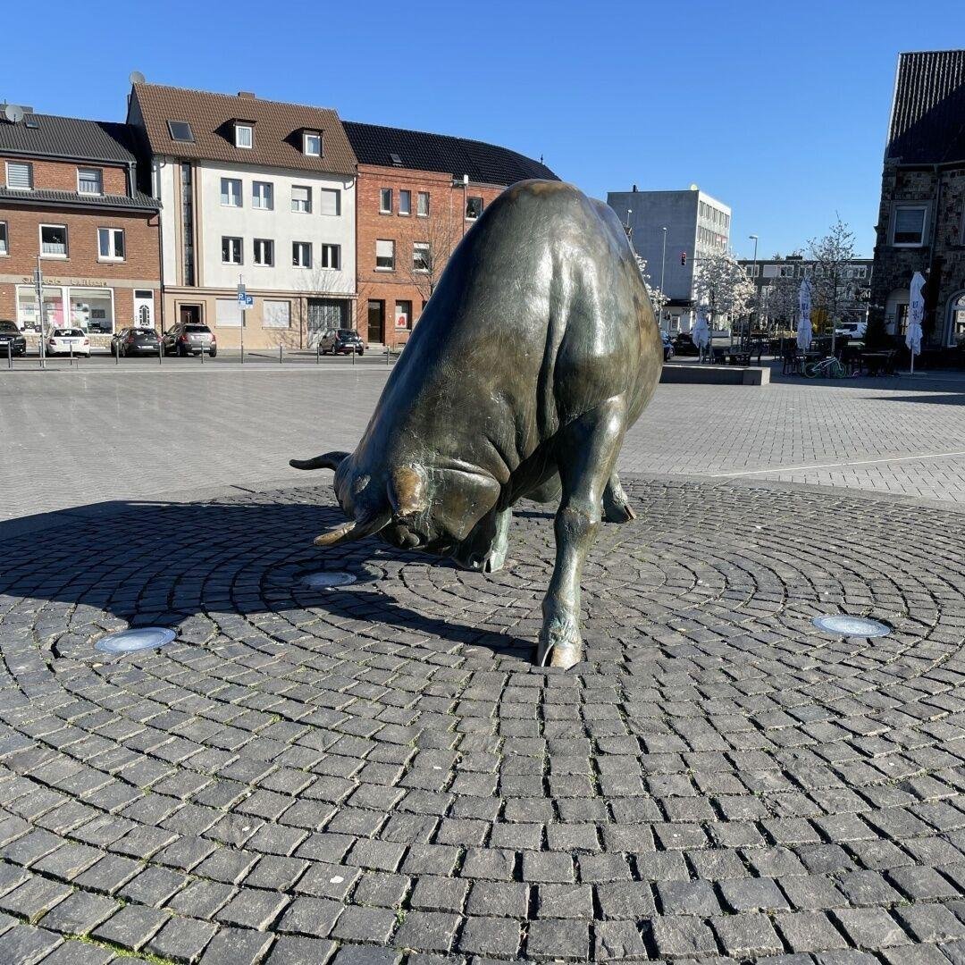 Eine lebensgroße Bronzestatue von einem Stier, der Kopf ist geneigt.
Im Hintergrund sieht man verschiedene Gebäude, der Himmel ist strahlend Blau.